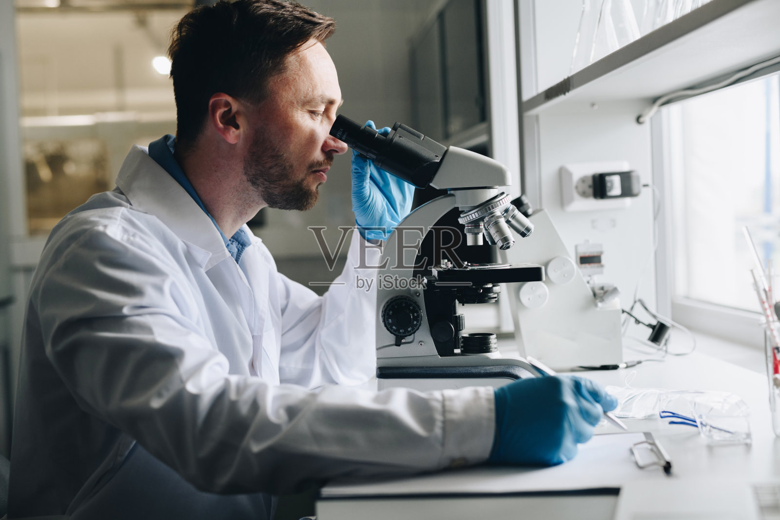 严肃专注的男性科学家穿着实验服，戴着一次性手套，在实验室里拿着研究:他坐在桌子旁，用显微镜专注于细胞照片摄影图片