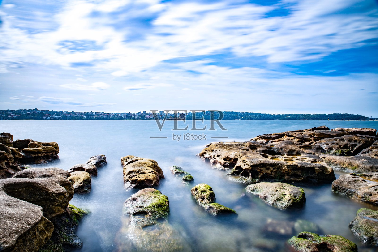 在一个多云的下午，长时间曝光在悉尼港，美丽的岩石在前景，柔软的海浪冲击海岸和美丽的海港前海岸为背景照片摄影图片