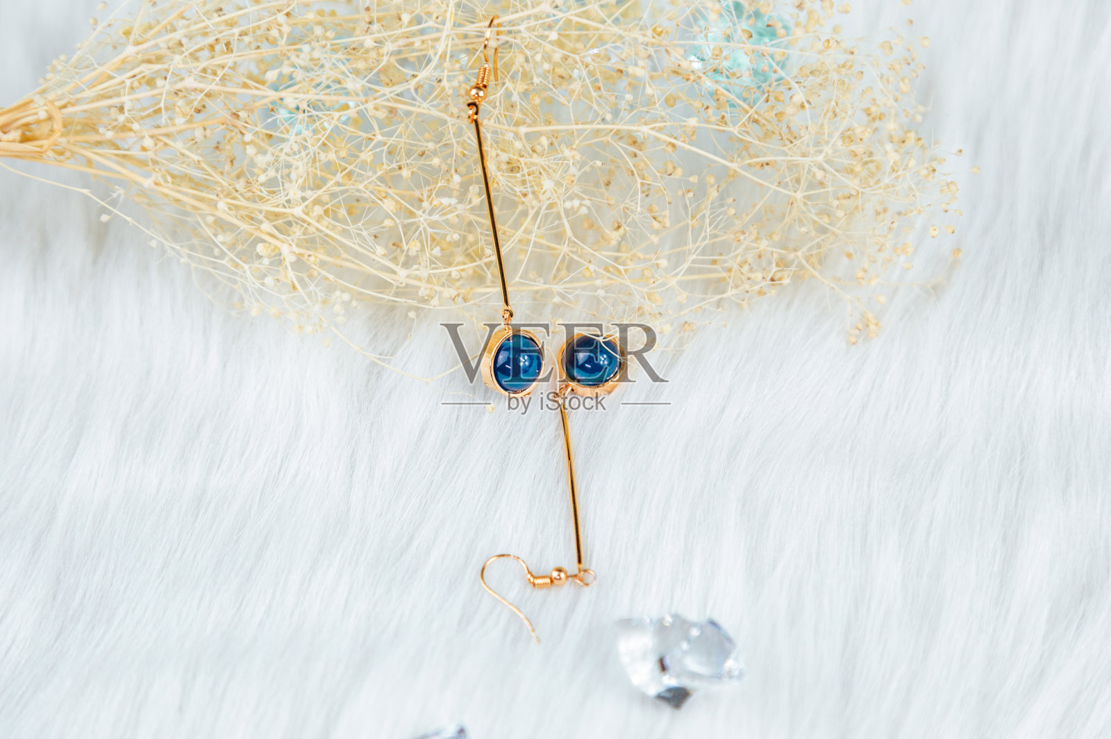 蓝色圆珠和金黄色金属制成的耳环。黄色干草植物作为背景。白色长毛绒布料和透明冰饰品。照片摄影图片