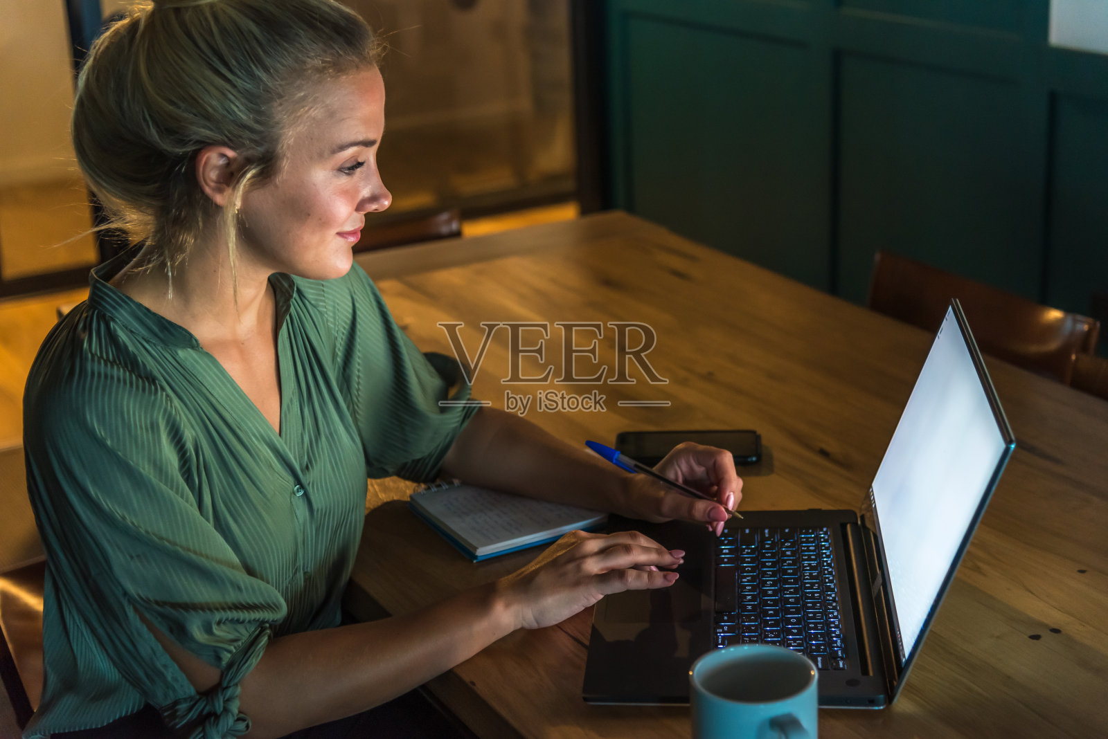 年轻的金发女人在她昏暗的笔记本前工作。桌子上有一个杯子，一个笔记本和一个手机照片摄影图片
