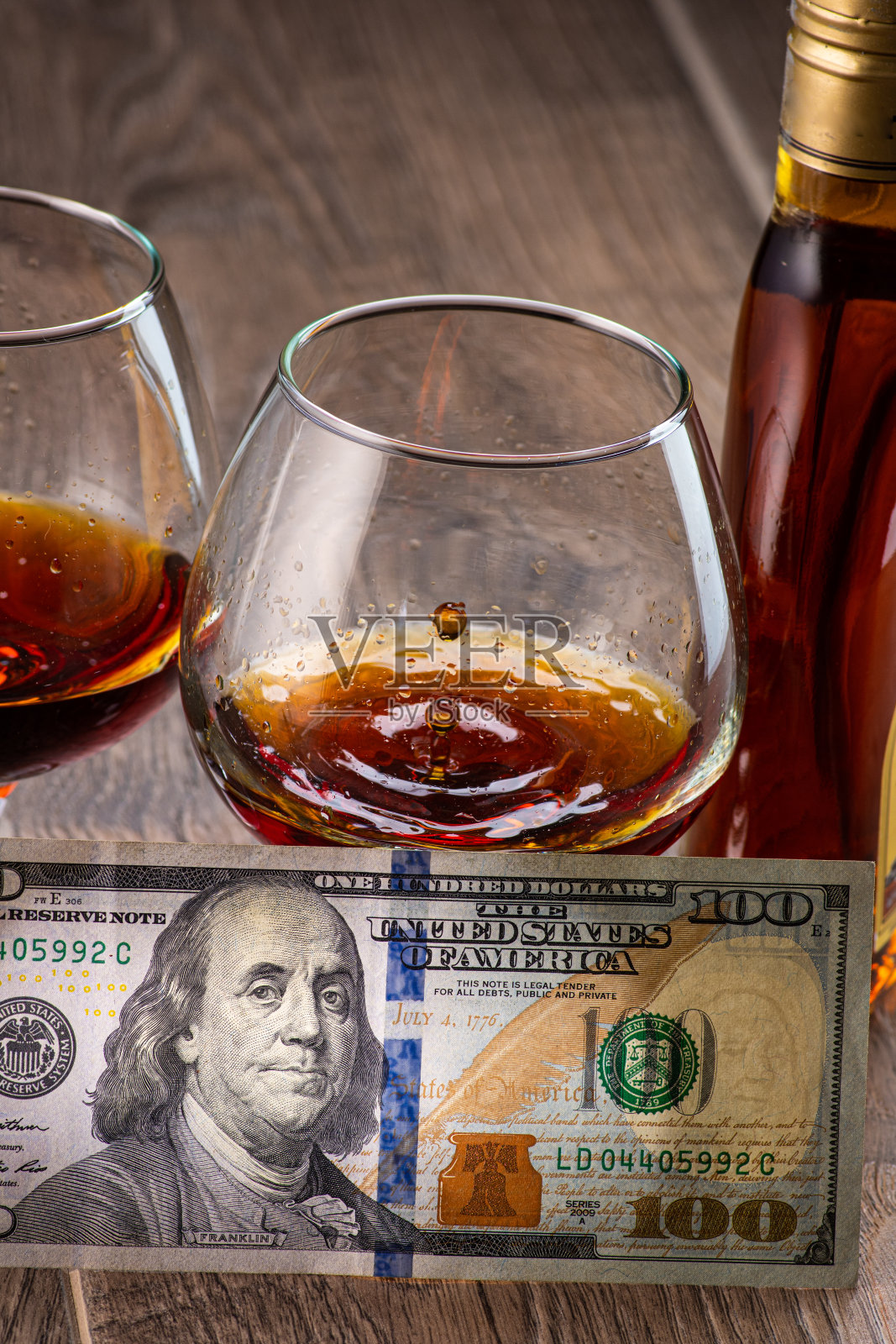 两杯干邑白兰地，在酒瓶旁边洒上一点，木背景上放着美元照片摄影图片