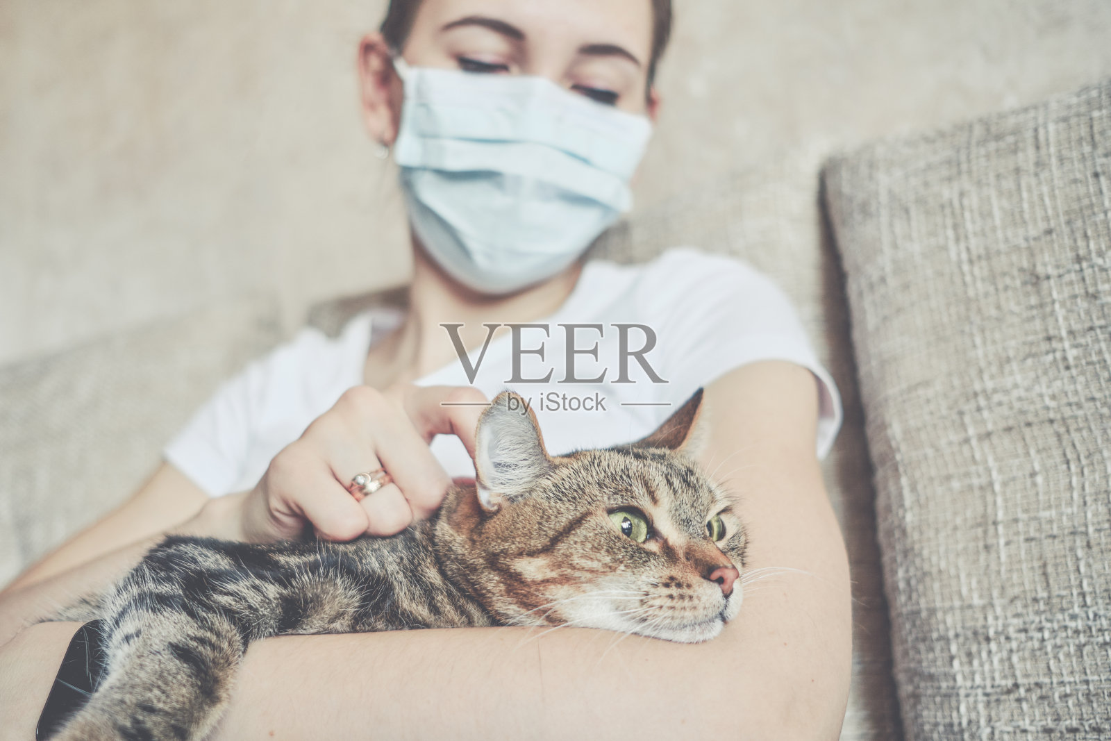 这个女孩戴着医用口罩在家自我隔离，和一只猫一起在沙发上休息。照片摄影图片