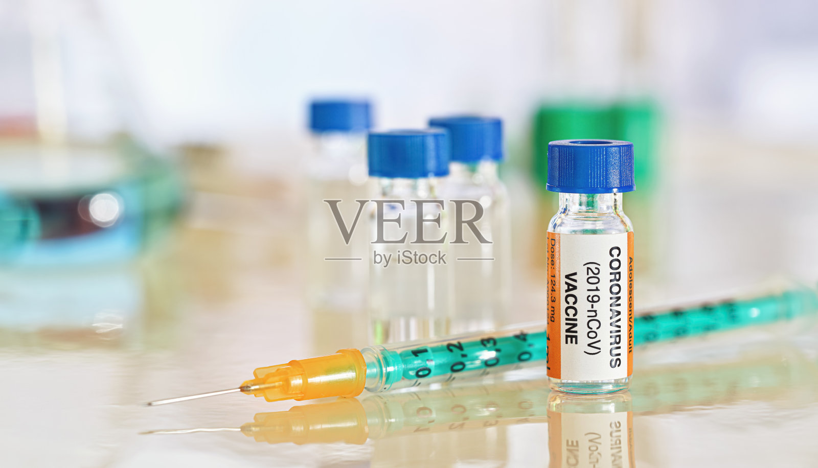 新冠肺炎疫苗概念——白色光滑的桌子上有蓝色盖子的小瓶子，旁边有绿橙色的注射器，更多的实验室玻璃模糊的背景(贴纸是自己设计的——不是真正的产品)照片摄影图片
