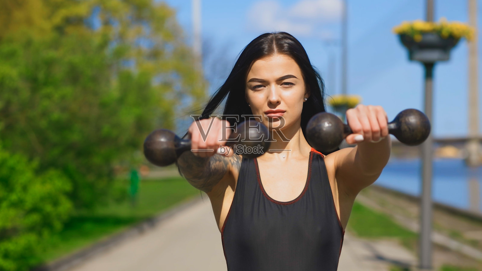 一个身材苗条的黑发女人在阳光明媚的日子里举着两个旧哑铃看着你照片摄影图片