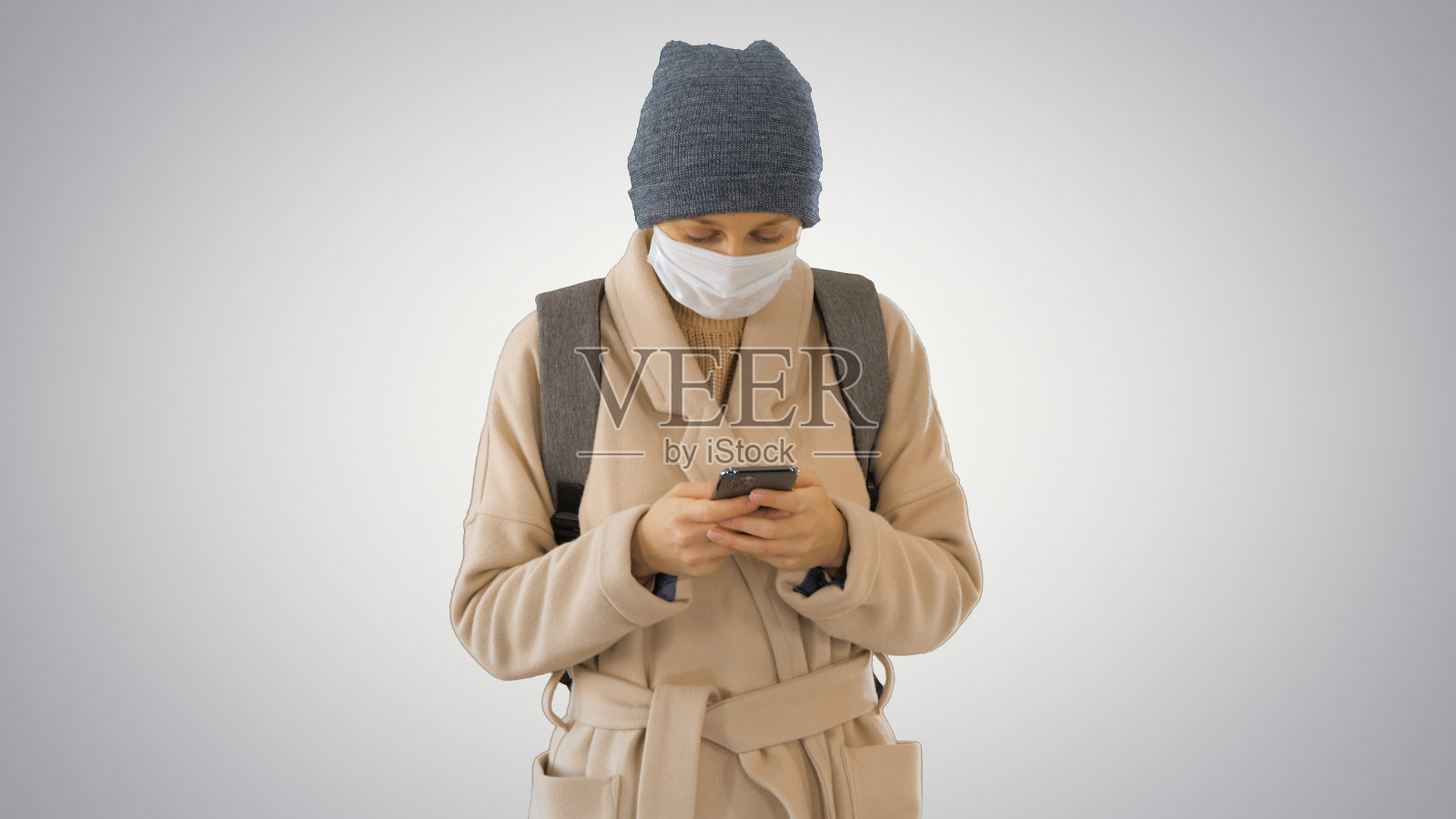 一名女子穿着一件大衣，戴着医用口罩，使用手机，在梯度背景上行走照片摄影图片