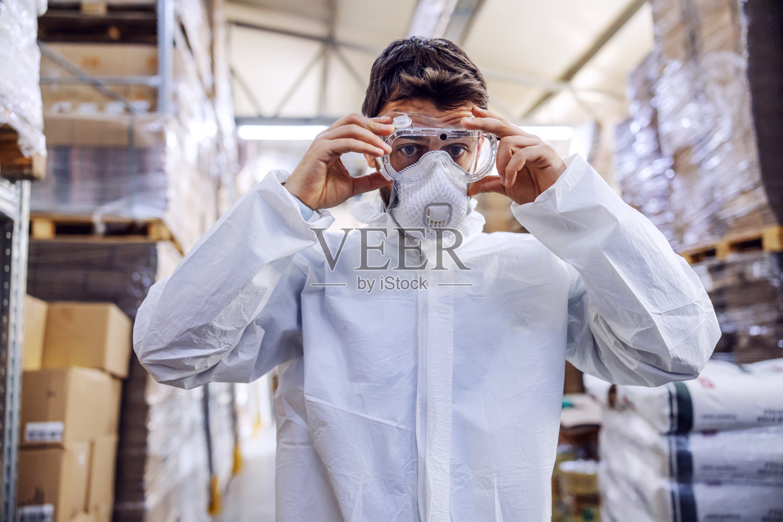 年轻男子身穿防护服，戴上防护眼镜，准备接受冠状病毒/ covid-19的消毒。仓库内部。仓库里全是食品。照片摄影图片