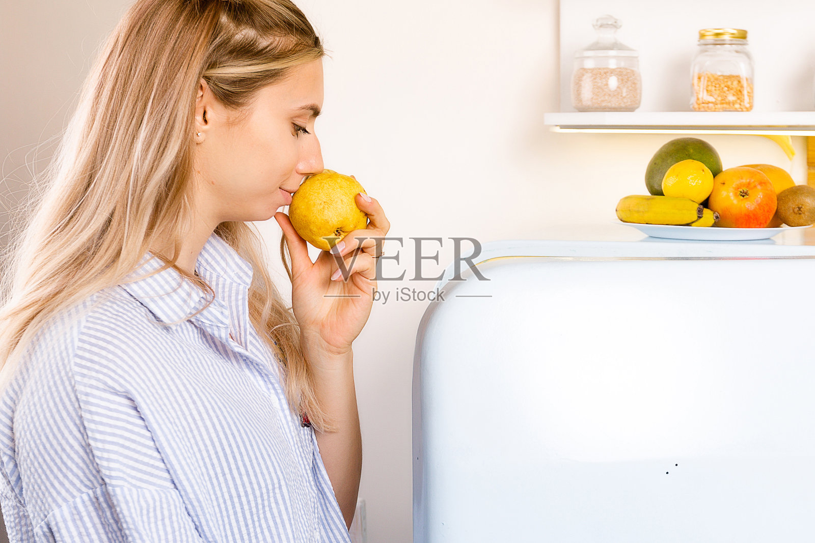 一个漂亮的年轻女人在厨房里拿着新鲜健康的食物。与copyspace概念。女孩在冰箱附近嗅和检查水果的新鲜程度照片摄影图片