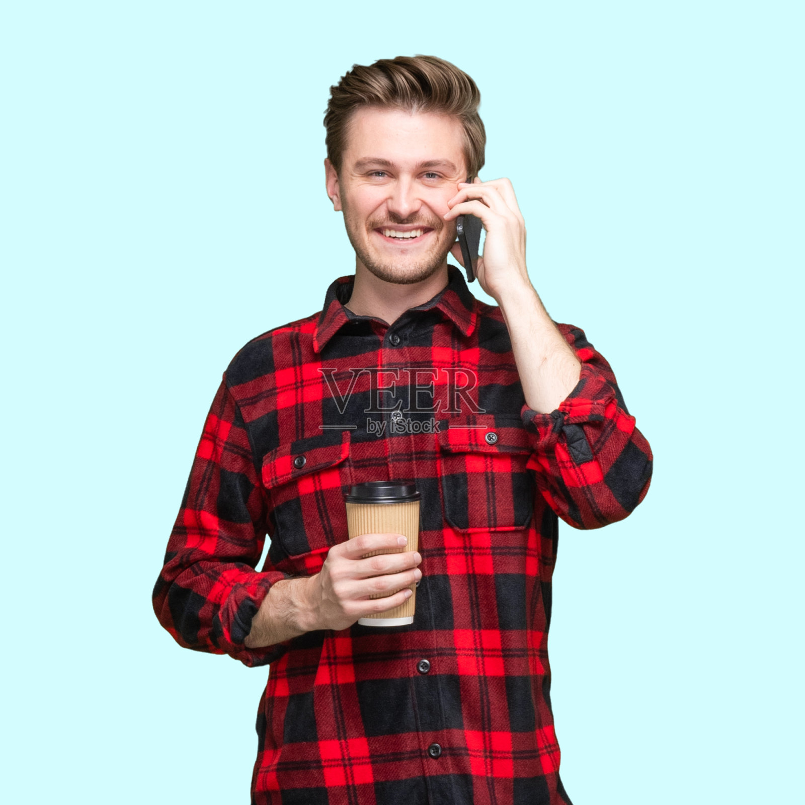白人年轻男性站在蓝色背景下，穿着纽扣衬衫，拿着咖啡杯和使用手机照片摄影图片