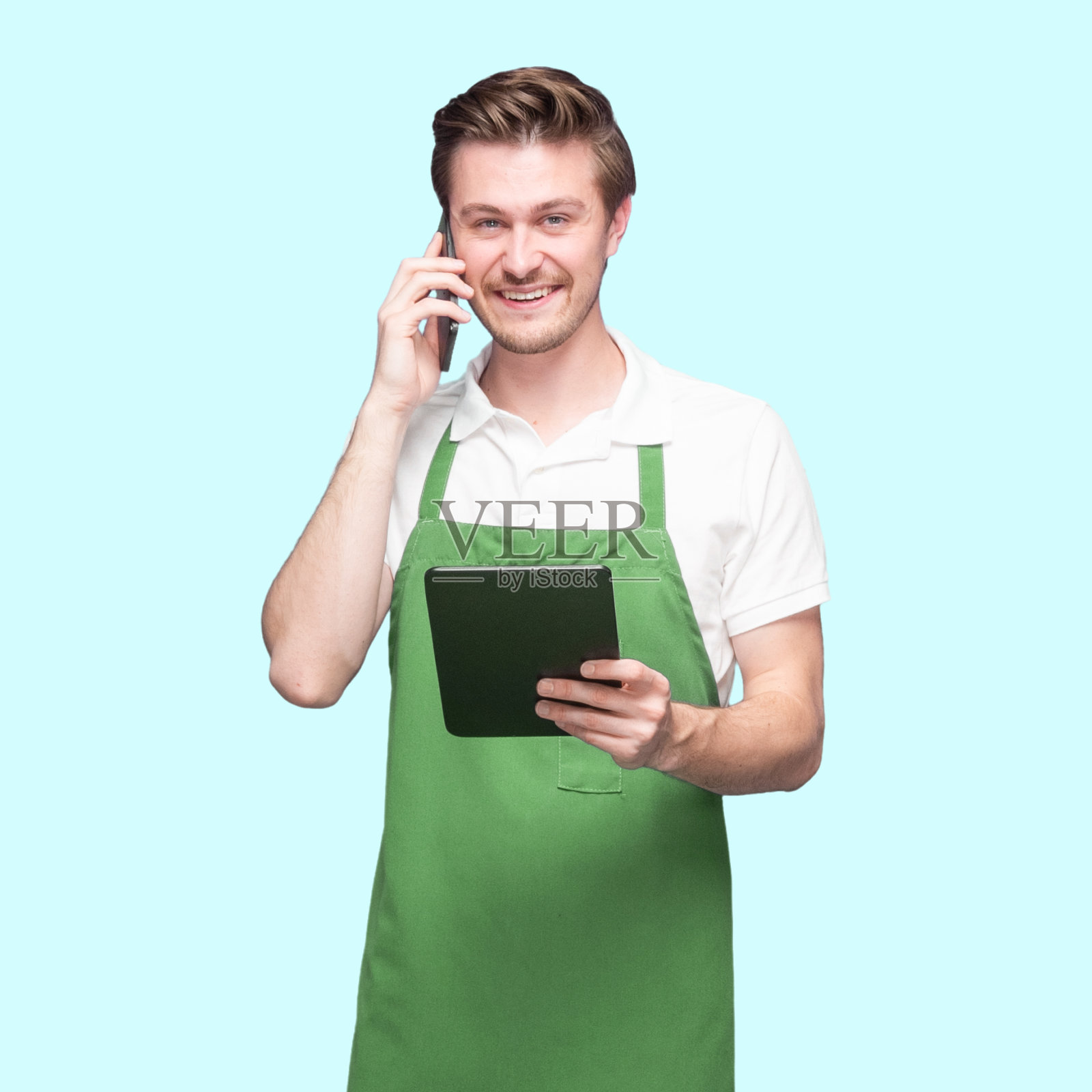 白人男咖啡师穿着围裙站在蓝色背景前，用着平板电脑照片摄影图片