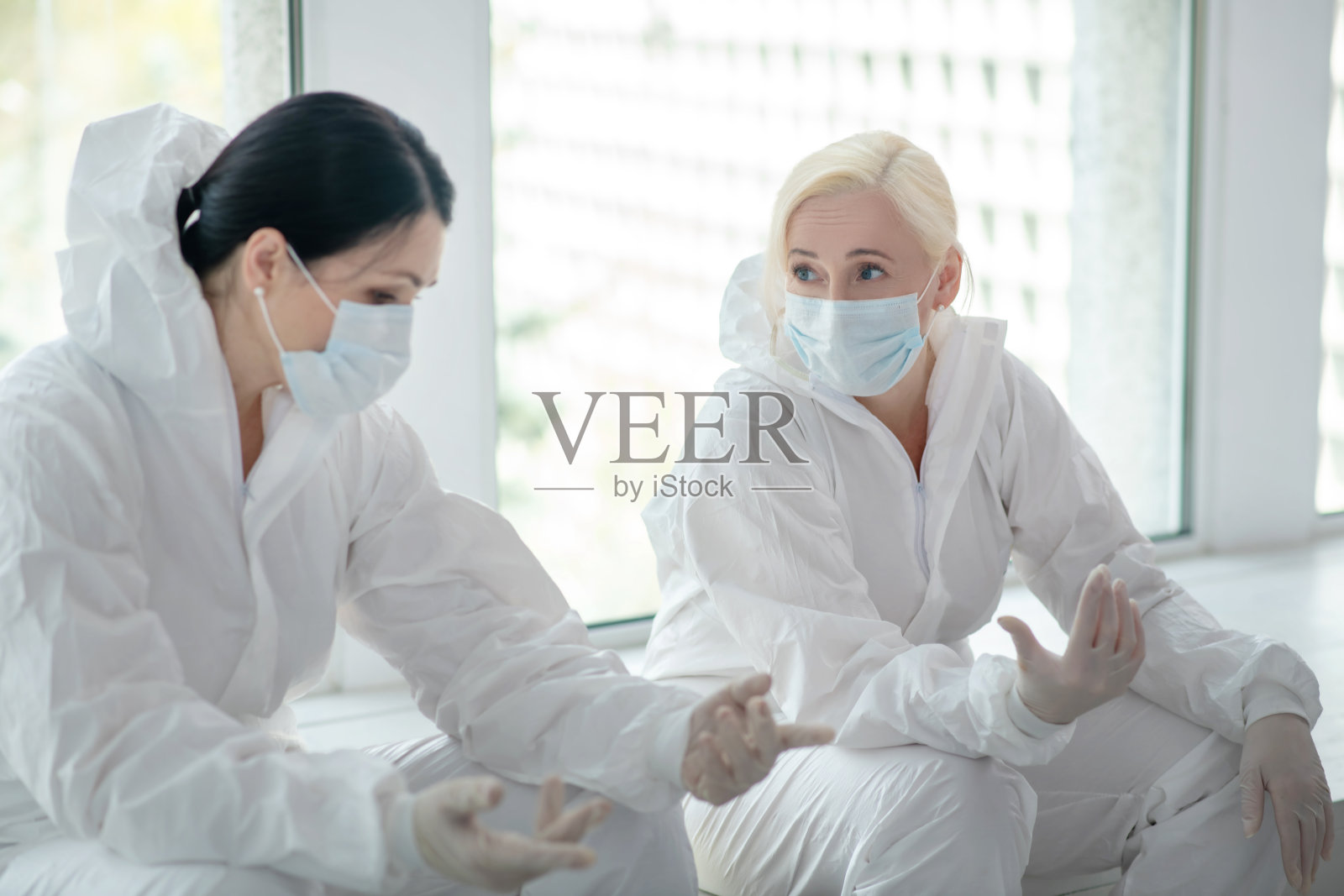 两名女医务人员穿着防护服，戴着医用口罩，坐在窗边聊天照片摄影图片