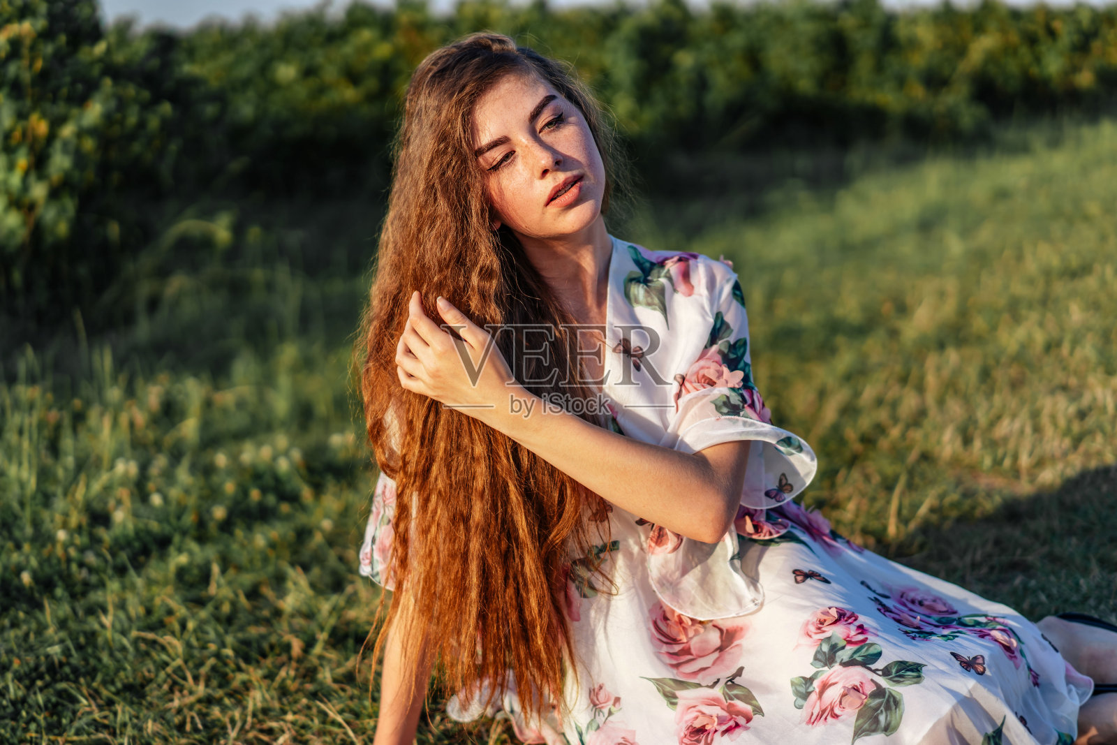 年轻的女孩有着长长的卷发，深色头发，脸上有雀斑，化着妆，有着绿色的眼睛，穿着浅白色的连衣裙在镜头前摆姿势，背景模糊，自然。照片摄影图片