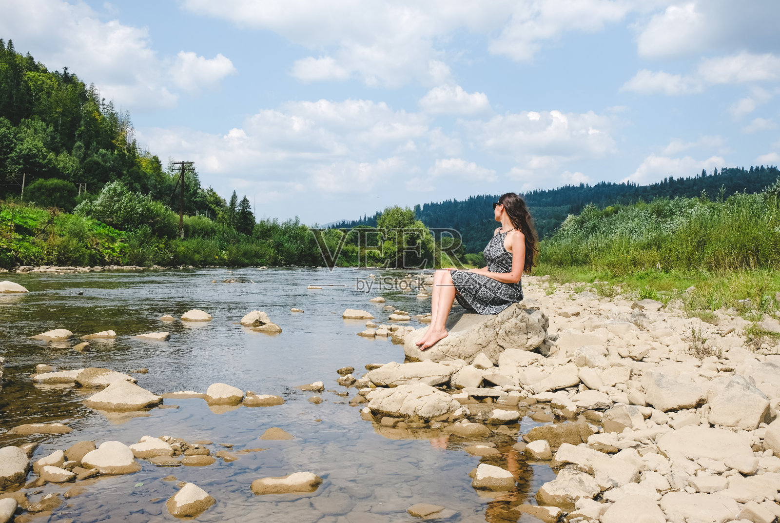 黑发女孩戴着墨镜坐在山河的石头上。后面是喀尔巴阡山脉。阳光明媚的夏天的天气。照片摄影图片