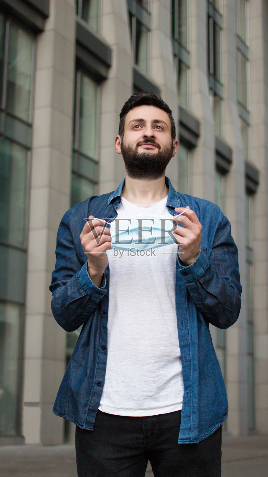 快乐的黑发男子身穿白色t恤和牛仔裤t恤，摘下医用口罩站在街道上，以办公楼为背景。结束隔离、covid-19大流行、自我隔离概念。照片摄影图片