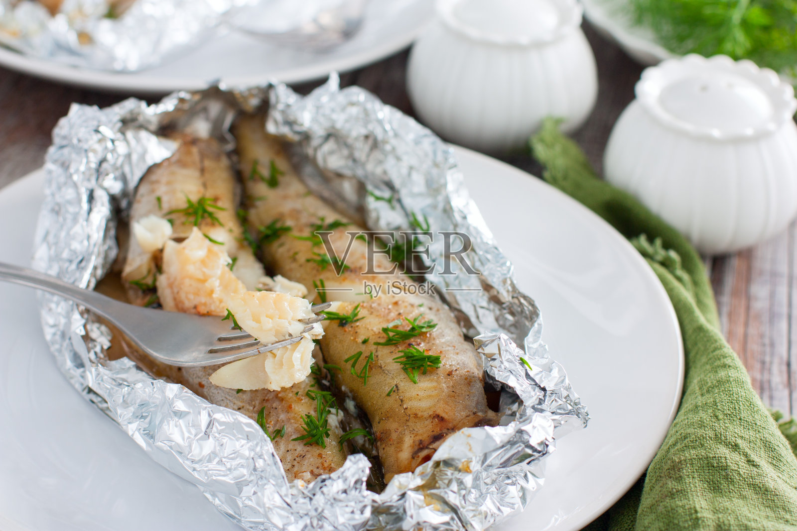 将整条鳕鱼用锡箔箔片放入烤箱中烘烤，盛于白色盘子上，选择性聚焦照片摄影图片