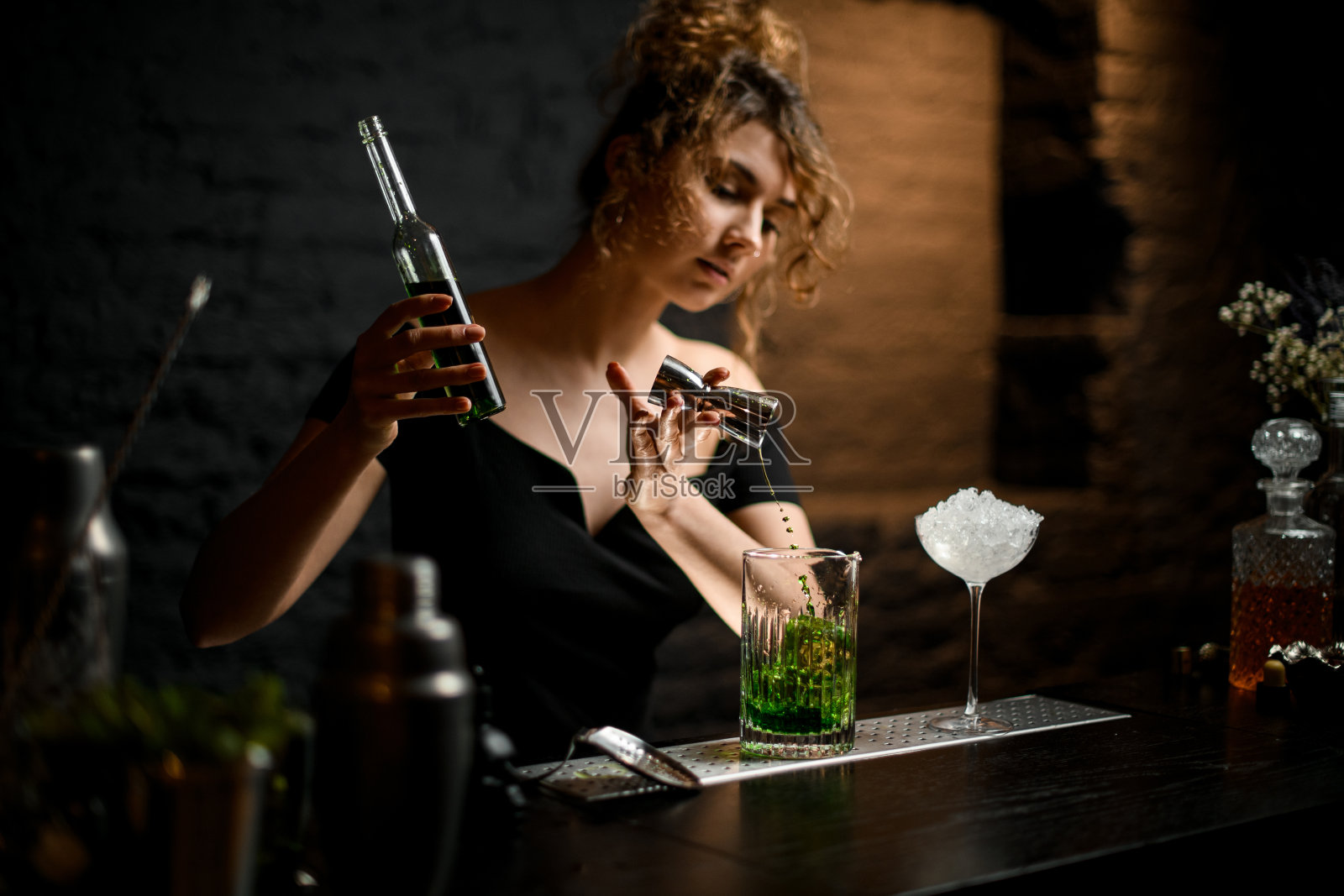 穿着黑衬衫的漂亮女孩酒保小心翼翼地将饮料从搅拌器倒入混合杯。照片摄影图片