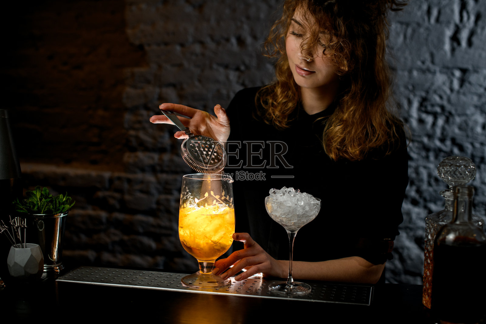 年轻的女酒吧招待拿着过滤网从杯子上盛着鲜亮的鸡尾酒。照片摄影图片