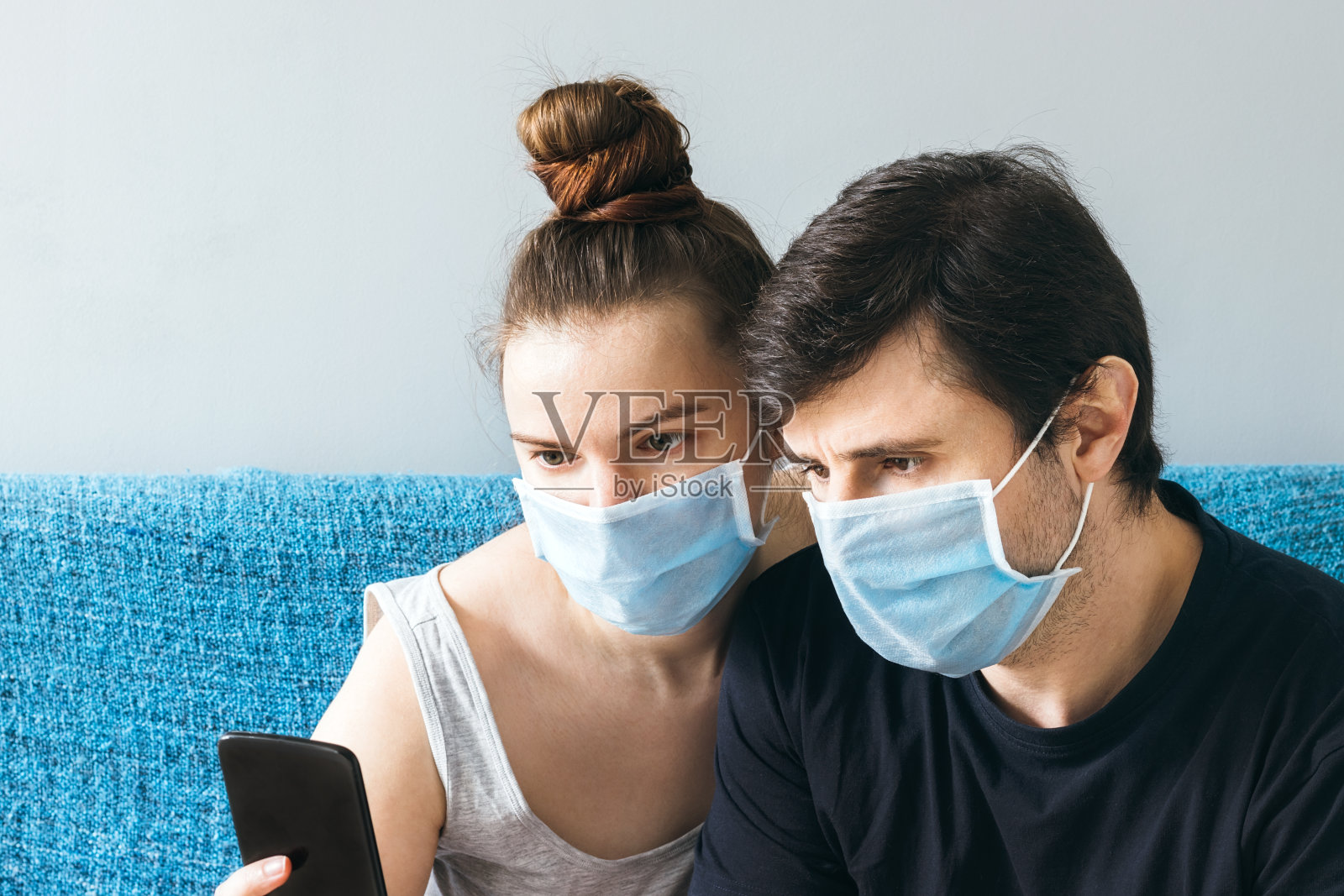 戴着蓝色医用口罩的夫妇在看智能手机上的东西。一男一女，一个两口之家，在隔离期间呆在家里照片摄影图片