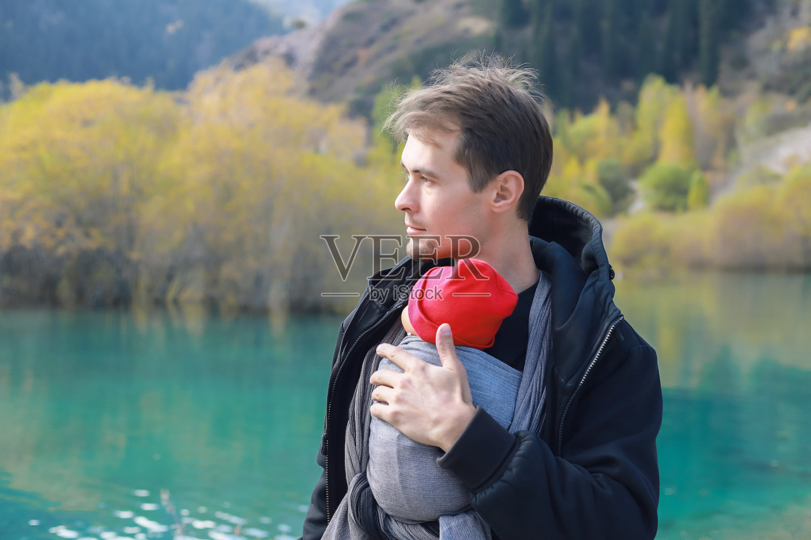 迷人的年轻父亲和她的婴儿在户外吊索。男人背着她的孩子，在秋天的山湖里旅行。婴儿袋的概念照片摄影图片