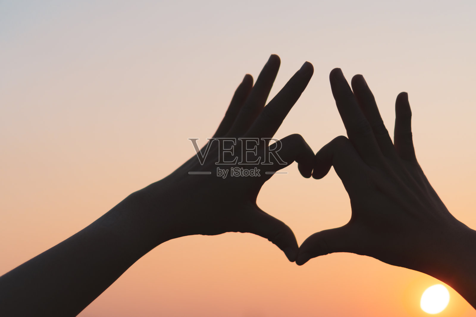 女人的手在夕阳的天空和散景的背景上做心形。照片摄影图片