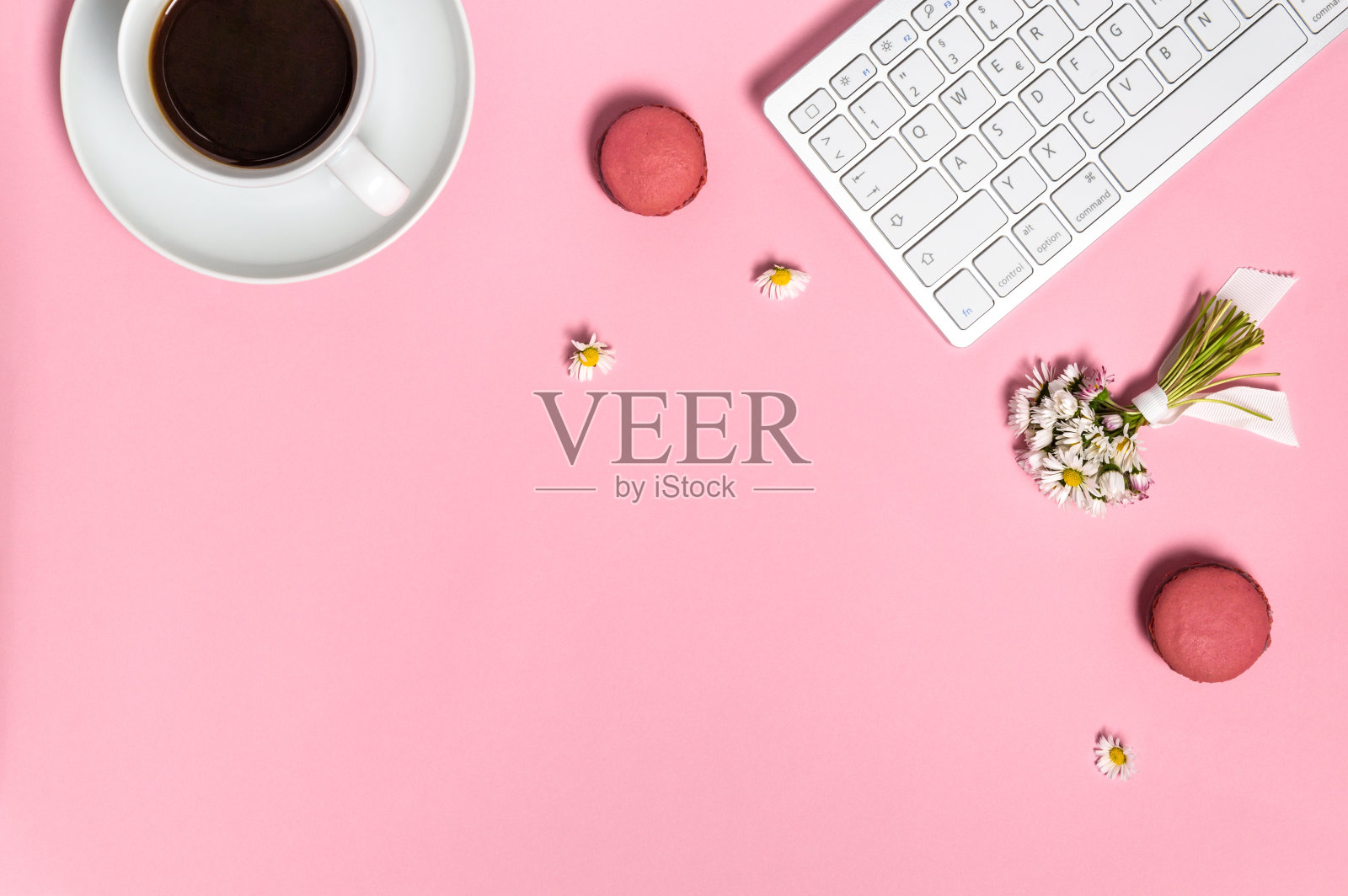 女性的工作空间有键盘，一杯黑咖啡，马卡龙和雏菊花在粉红色的背景，俯视图照片摄影图片