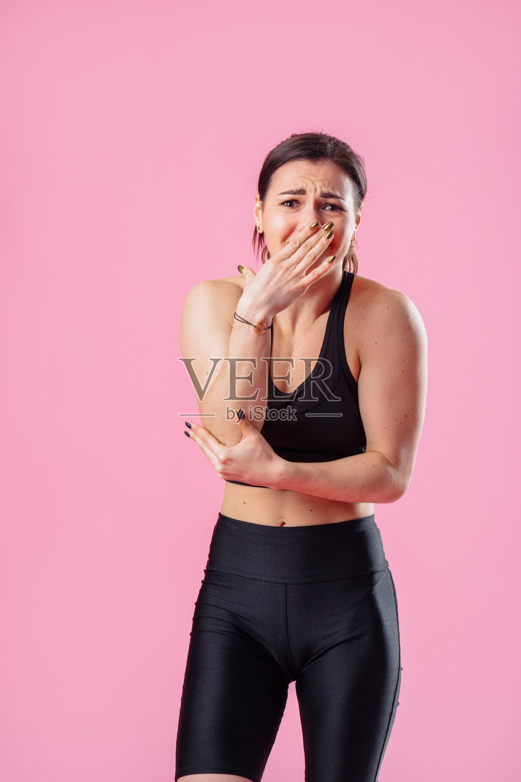 黑发女子与奇怪的绿色眼睛在黑色运动衫与震惊的脸关闭掩盖嘴与她的手在粉红色的墙壁背景照片摄影图片