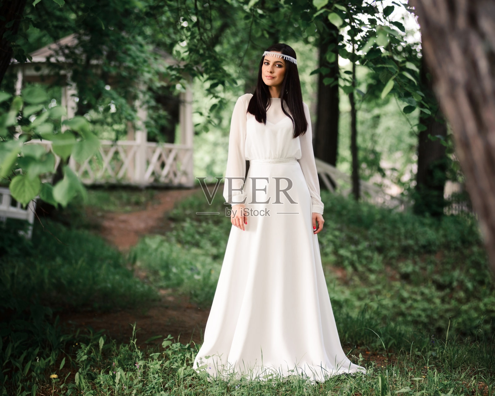 新娘身着优雅的婚纱，在一个夏日的绿色花园中，背景是一个古老的白色凉亭照片摄影图片