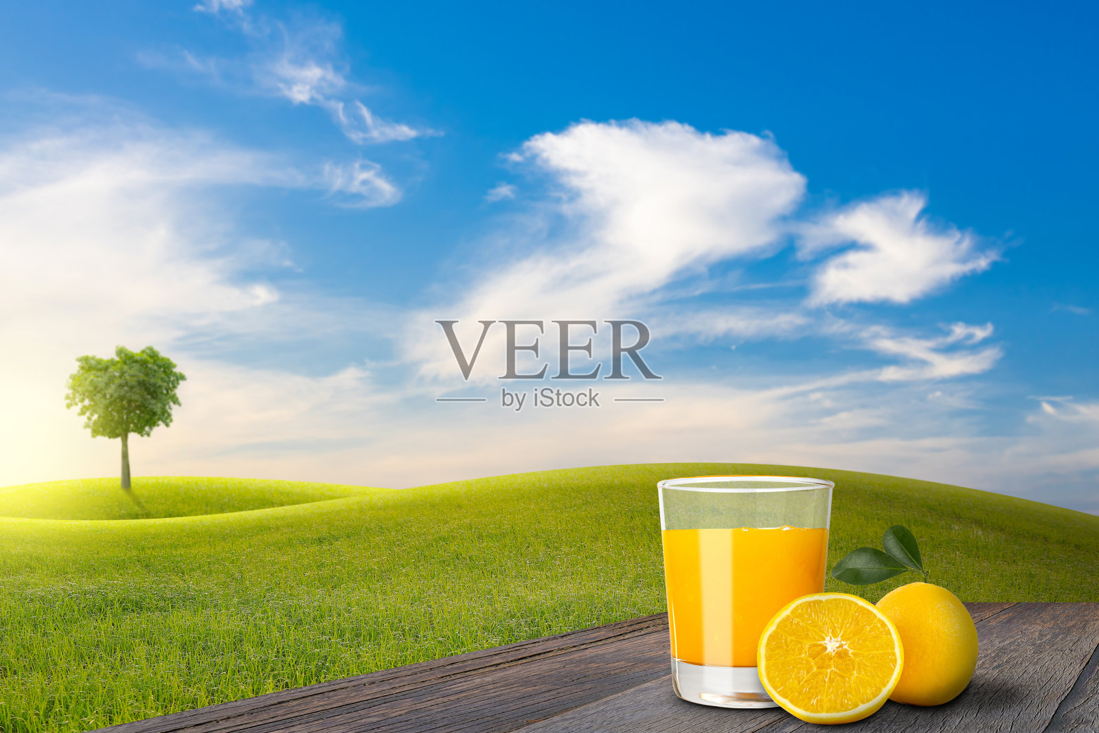 在破旧的木地板上，一杯橙汁和橘子，旁边是绿色的田野，山坡和树与蓝天和云彩的背景。照片摄影图片