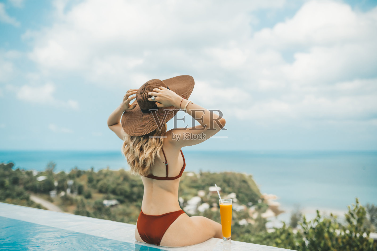 戴着帽子的金发女人微笑着坐在游泳池边喝着芒果奶昔照片摄影图片