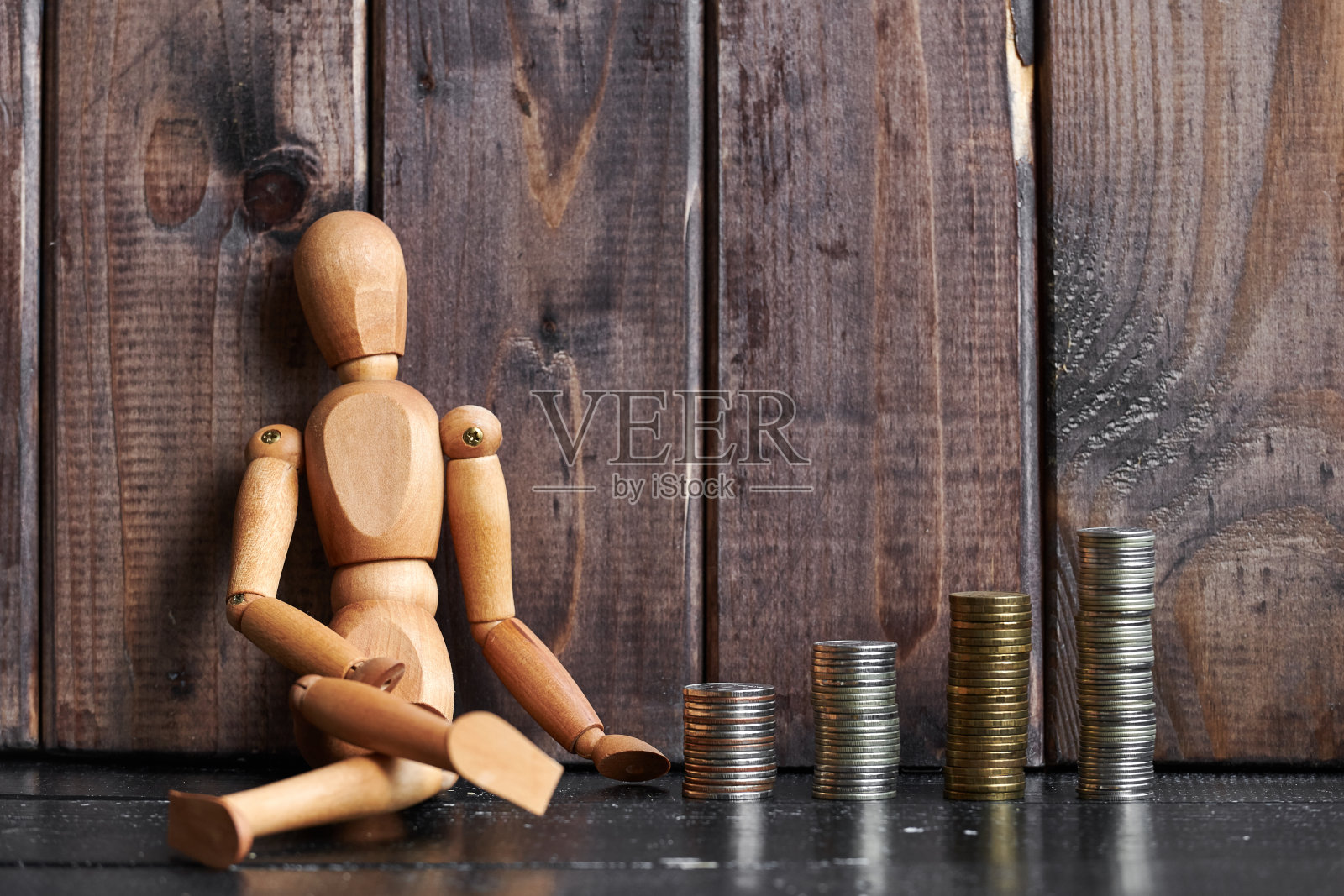 一个木制的男人模型坐在成堆的钱旁边，木质的背景照片摄影图片
