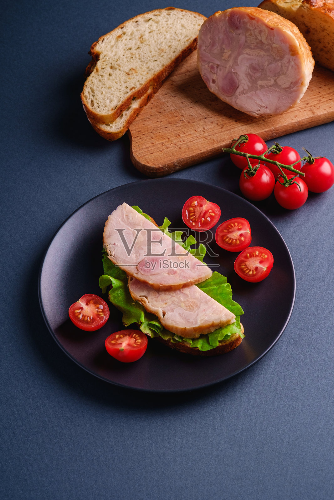 夹有火鸡火腿肉、青菜沙拉和新鲜樱桃番茄片的三明治放在黑色的盘子里，靠近砧板上的食材，蓝色最小的背景，视角照片摄影图片