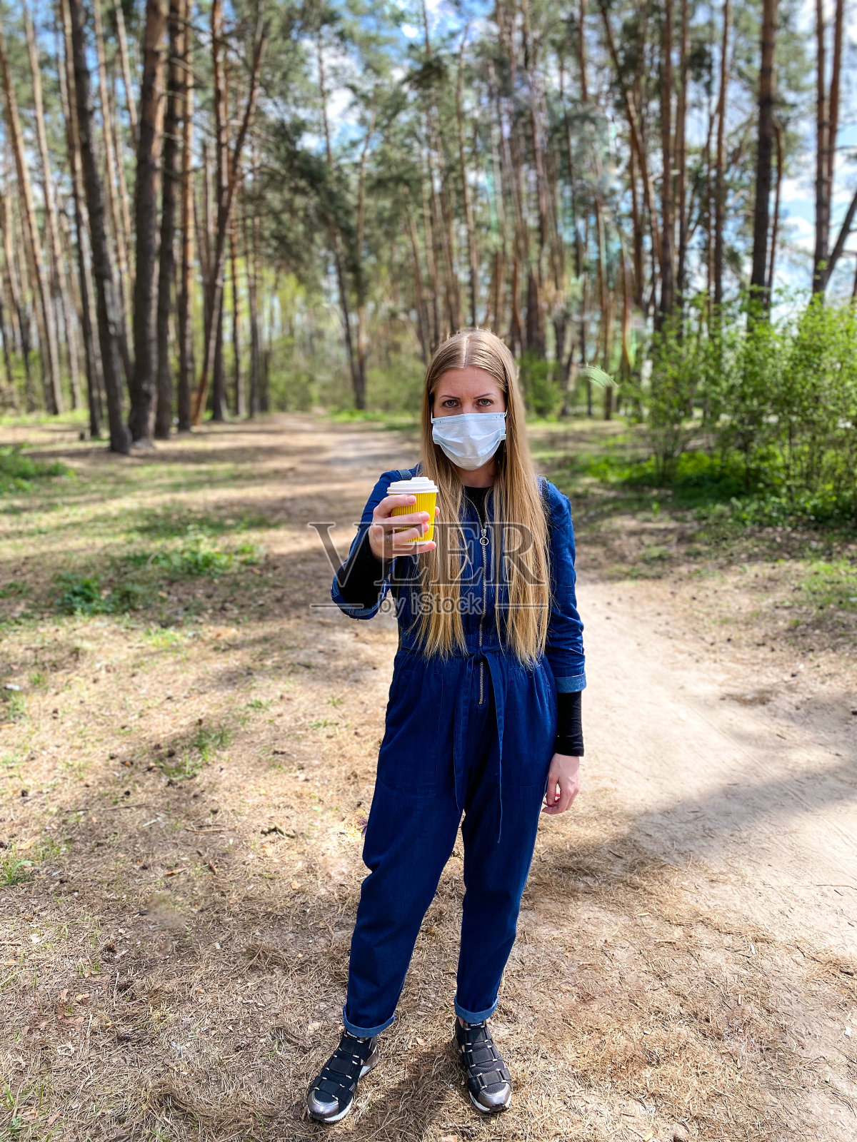 皮肤白皙的年轻女子/女孩，金色长发，带着医用防护口罩，在春天的户外散步时，手持一杯咖啡/热饮。照片摄影图片