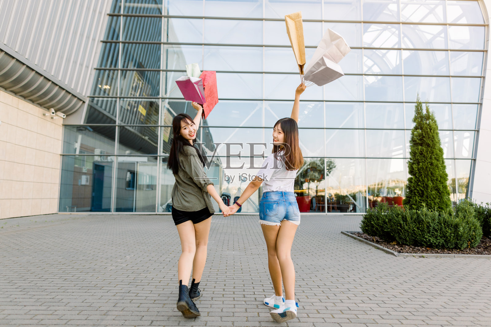 年轻迷人的中国女孩最好的朋友穿着休闲时尚的衣服，手牵着手，拿着购物袋一起走在现代化的城市购物中心前。照片摄影图片
