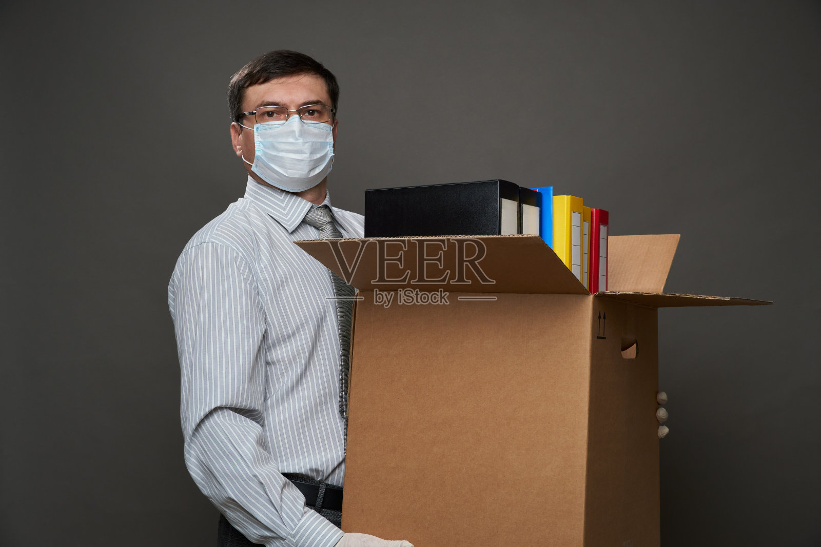一名打扮成商人的男子拿着一个装满办公用品、文件的箱子，在灰色背景下摆姿势，戴着医用口罩、防护手套、眼镜、衬衫和领带照片摄影图片