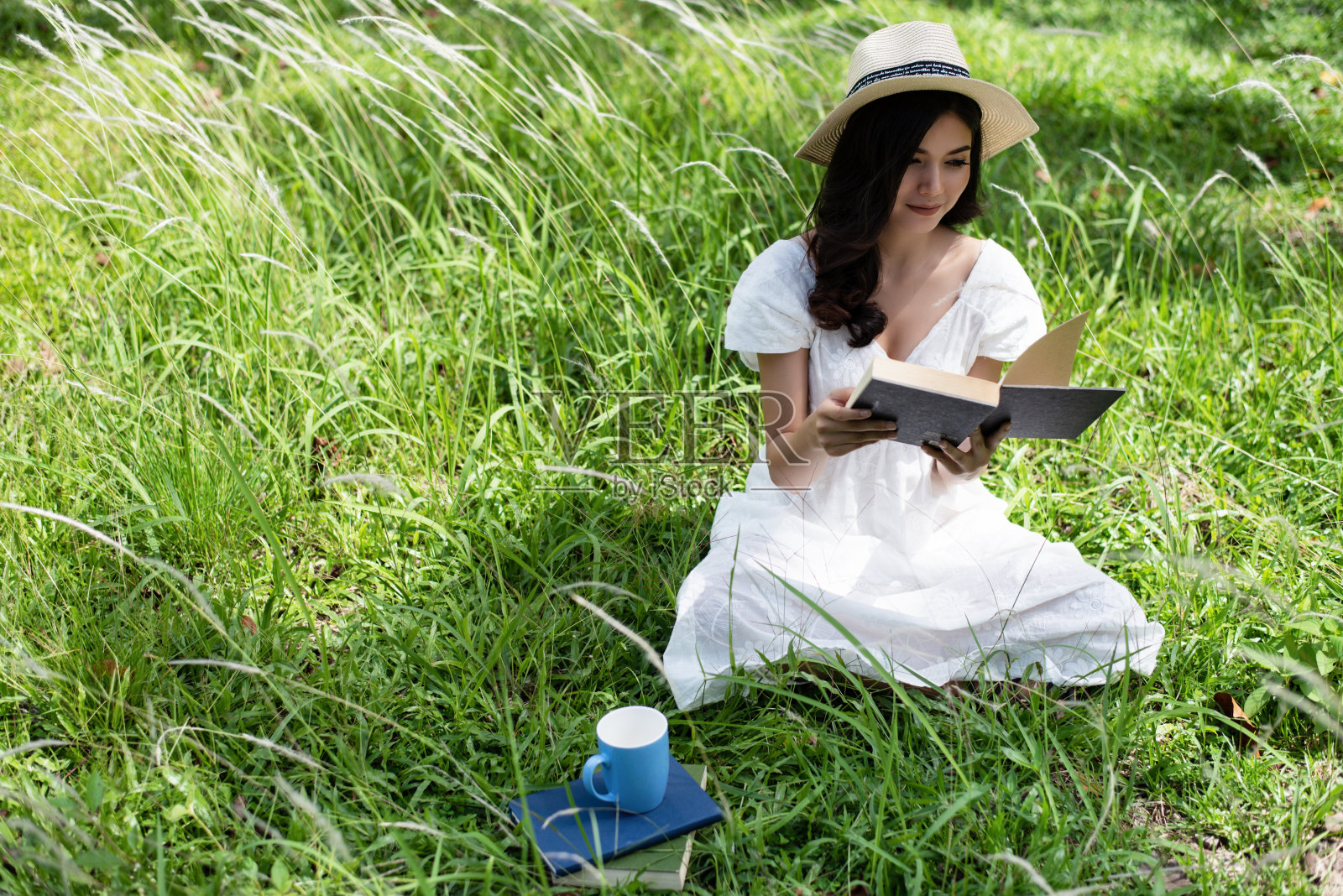 穿着白衣的美女在花园里，坐在绿色的草地上，看书，旁边是陶瓷咖啡杯，在花园里，朦胧的灯光周围。照片摄影图片