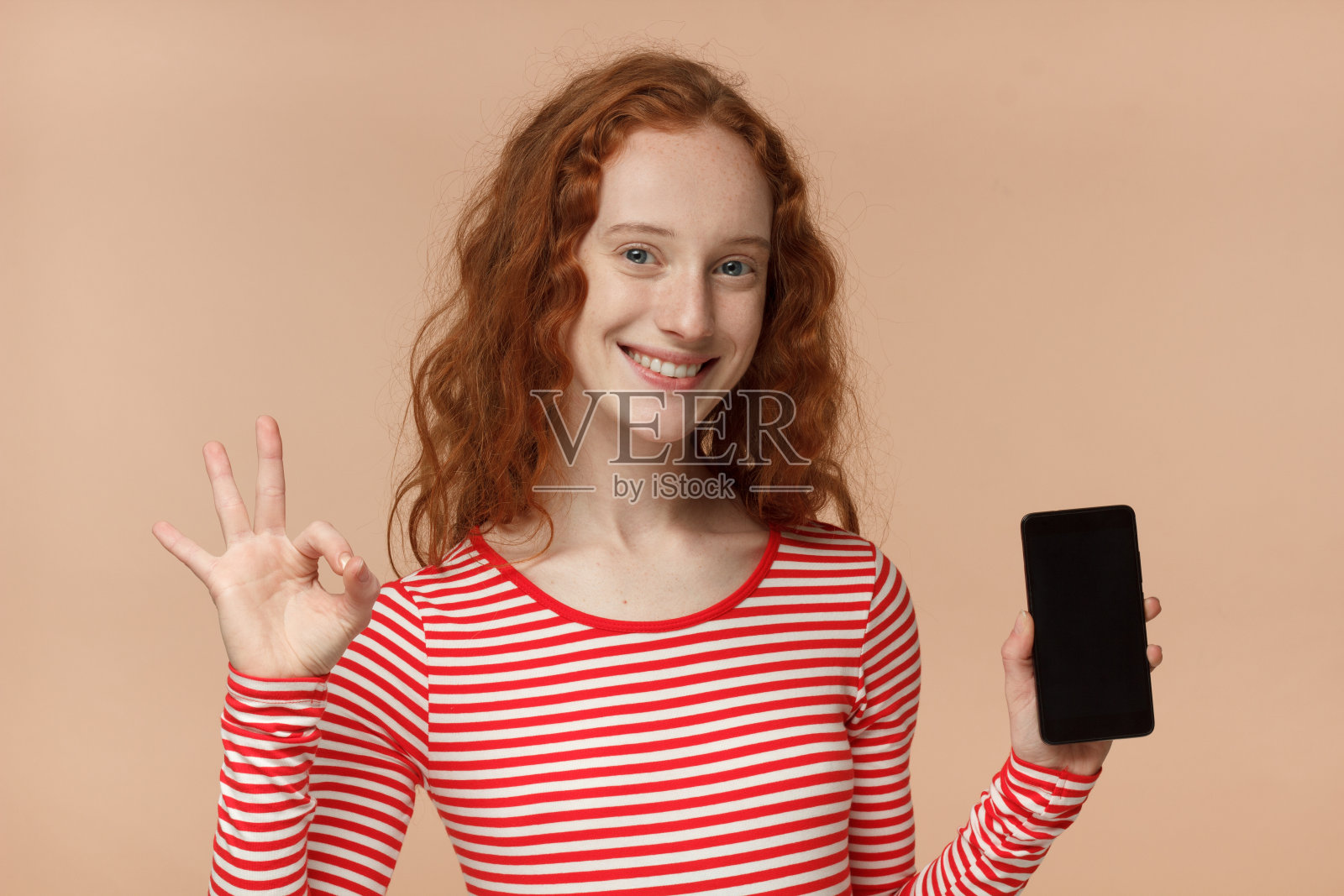 室内肖像迷人的漂亮的卷发红发女孩，拿着空白的智能手机，对着相机微笑，显示好的姿态，感到高兴照片摄影图片