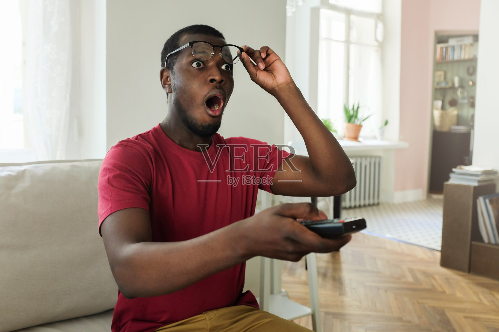 水平图片的年轻非洲男子花空闲时间在家里看电视，看着惊讶和震惊与张开的嘴举起眼镜，以确保他看到的屏幕是真实的照片摄影图片