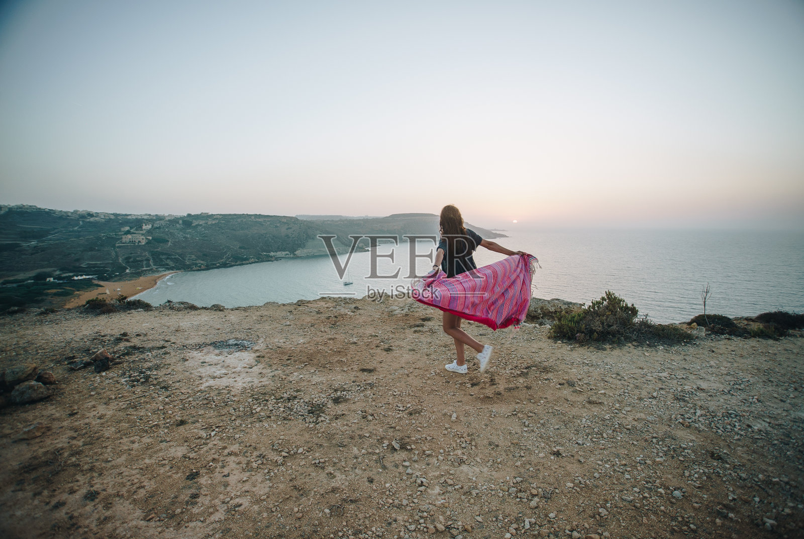 美丽的黑发旅行者女人跑与粉红色毯子卡利普索洞穴与美丽的拉姆拉湾与黄色的沙滩和地中海。Gozo、马耳他照片摄影图片