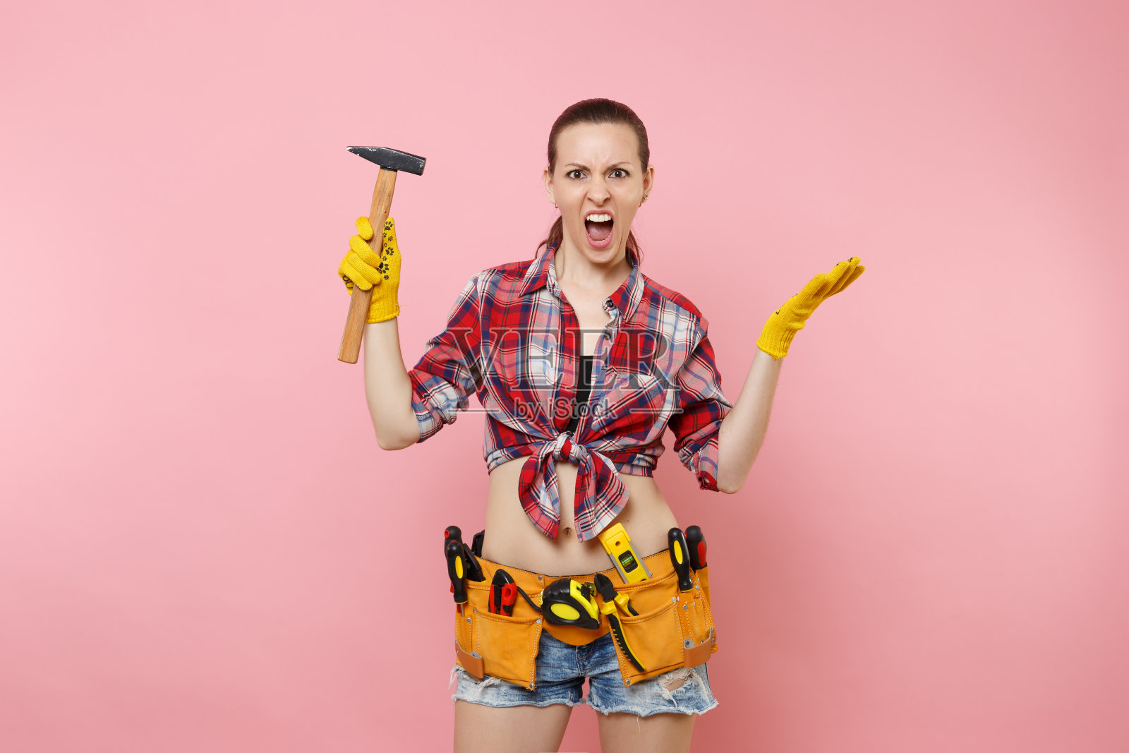 强壮的年轻杂工女人穿着格子衬衫，牛仔短裤，工具包工具皮带上满是不同的乐器锤孤立在粉红色的背景。女性从事男性的工作。装修和职业理念。照片摄影图片