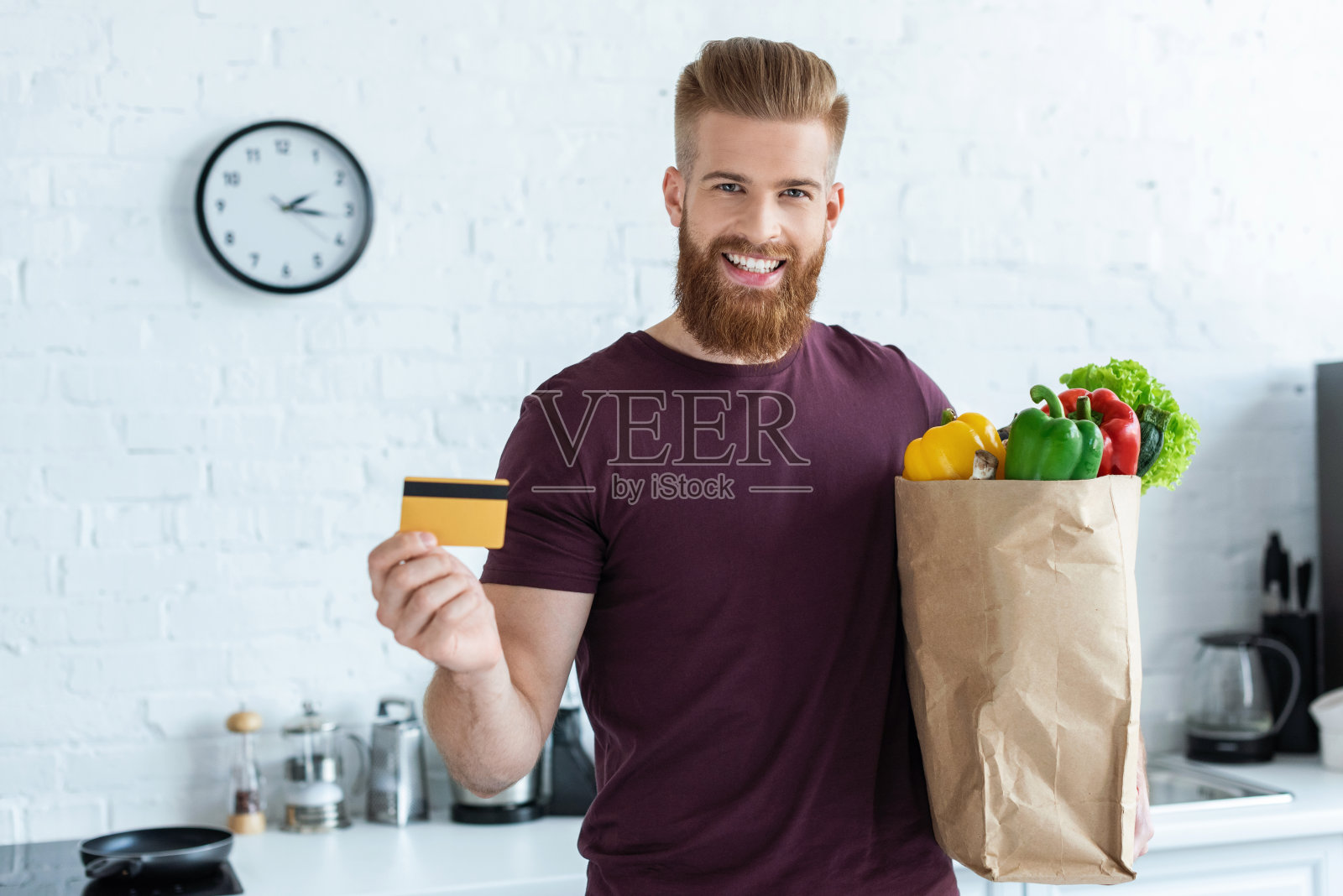 一个长着胡须的帅哥，拿着装着蔬菜和信用卡的购物袋，在厨房对着镜头微笑照片摄影图片