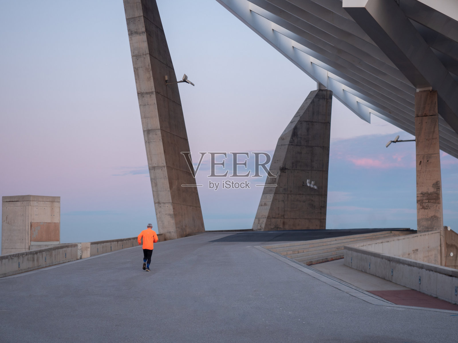 一个穿着橘色夹克的老人在日落时分在公园里跑步。(西班牙加泰罗尼亚巴塞罗那论坛公园)照片摄影图片