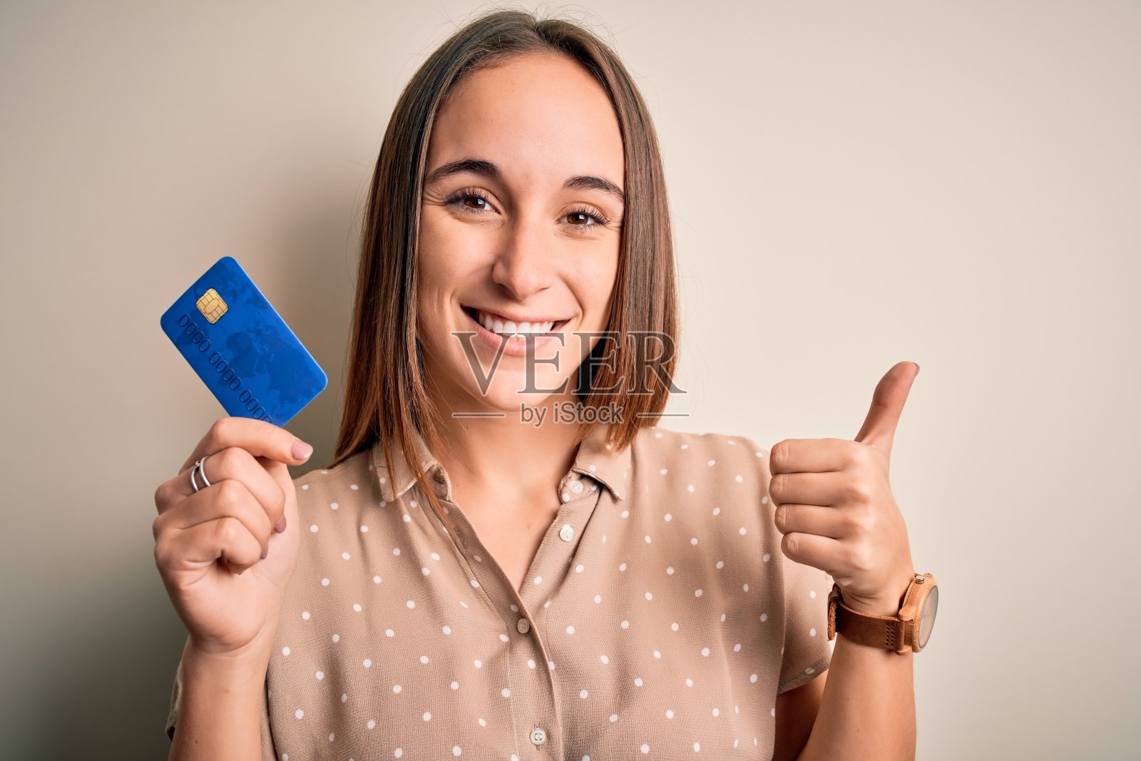 年轻美丽的顾客女人拿着信用卡做付款在白色的背景愉快地与大微笑做ok手势，拇指向上的手指，优秀的手势照片摄影图片