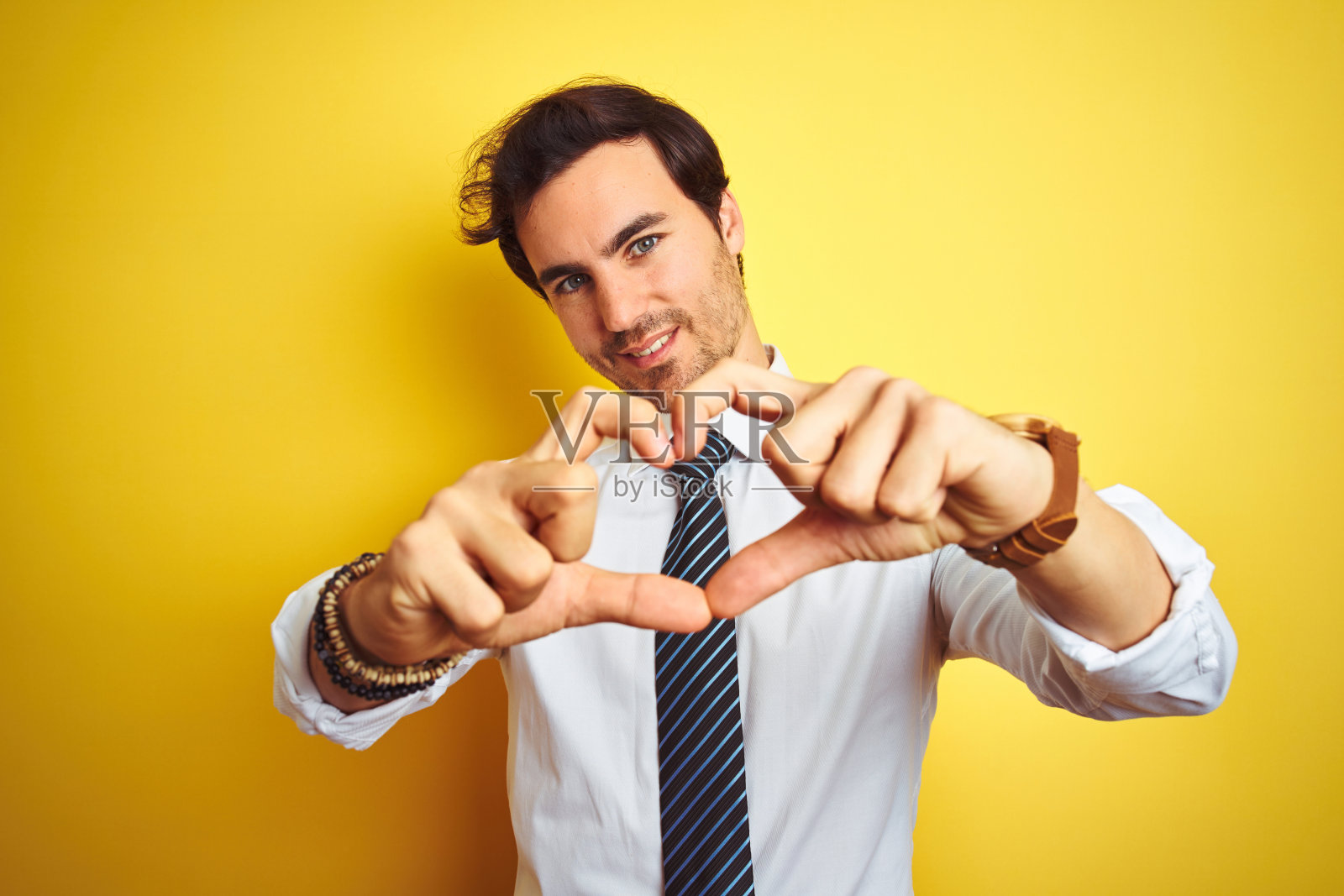 年轻英俊的商人穿着优雅的衬衫和领带在孤立的黄色背景微笑着做心形符号形状的手。浪漫的概念。照片摄影图片