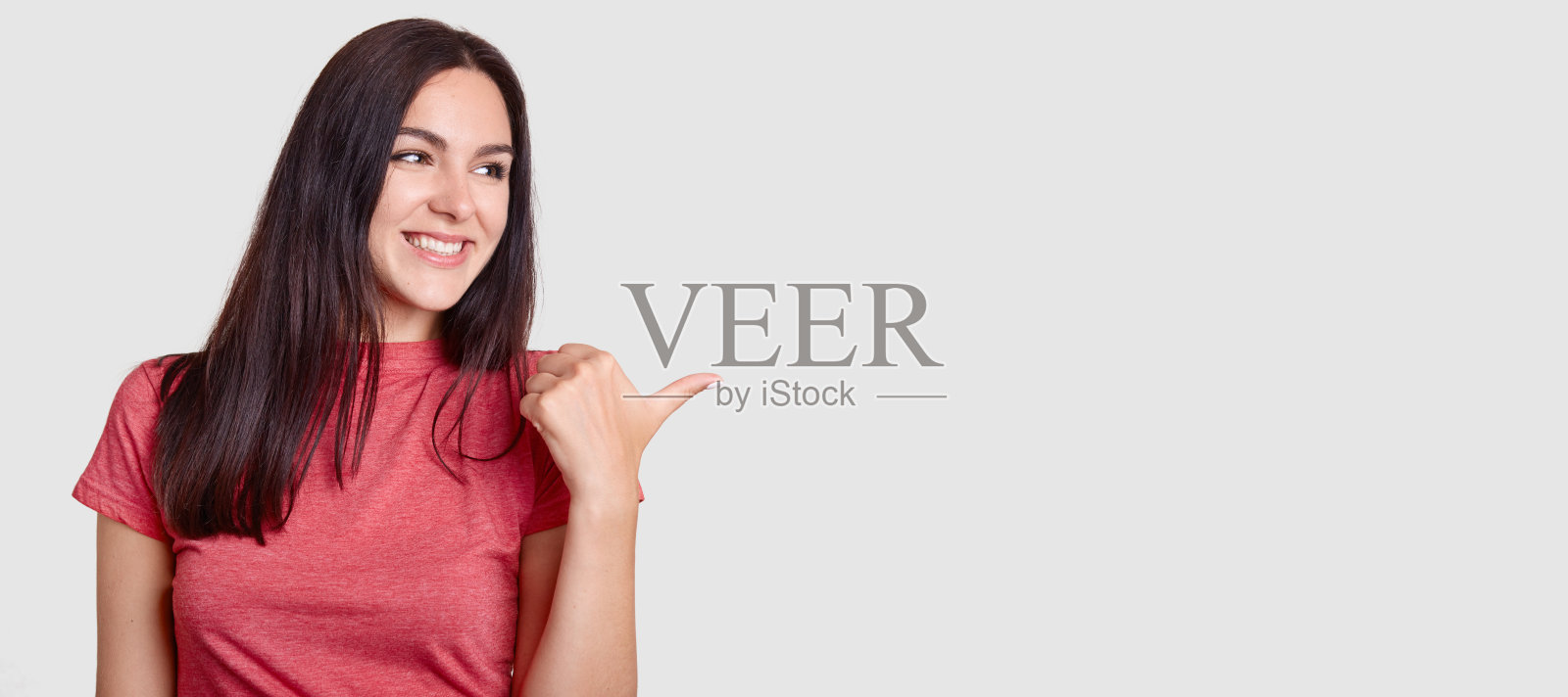 工作室拍摄的快乐的黑发女人，笑容灿烂，皮肤健康，参与广告活动，用拇指表示，穿着休闲t恤，显示您的广告空间照片摄影图片