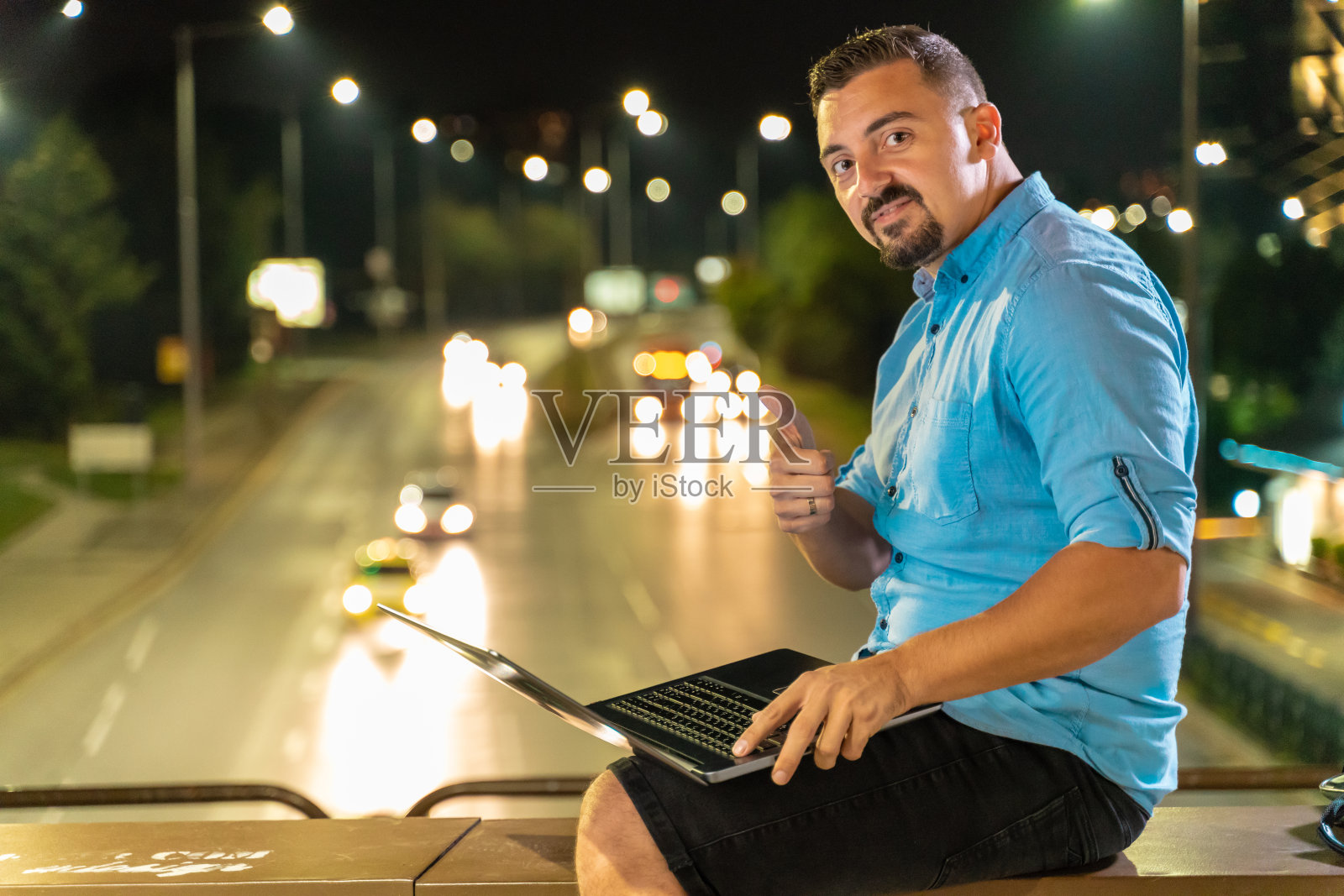 一名男子看着摄像头，微笑着竖起大拇指，晚上在户外拿着笔记本电脑照片摄影图片