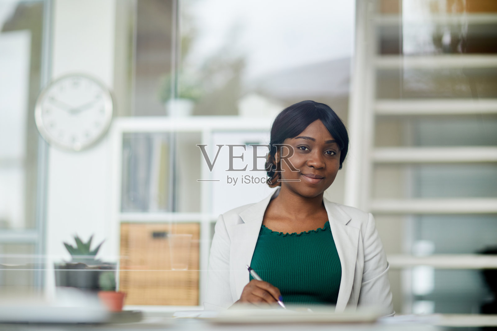 内容有吸引力的黑色女士在夹克坐在桌子上，看着相机，而在现代办公室工作照片摄影图片