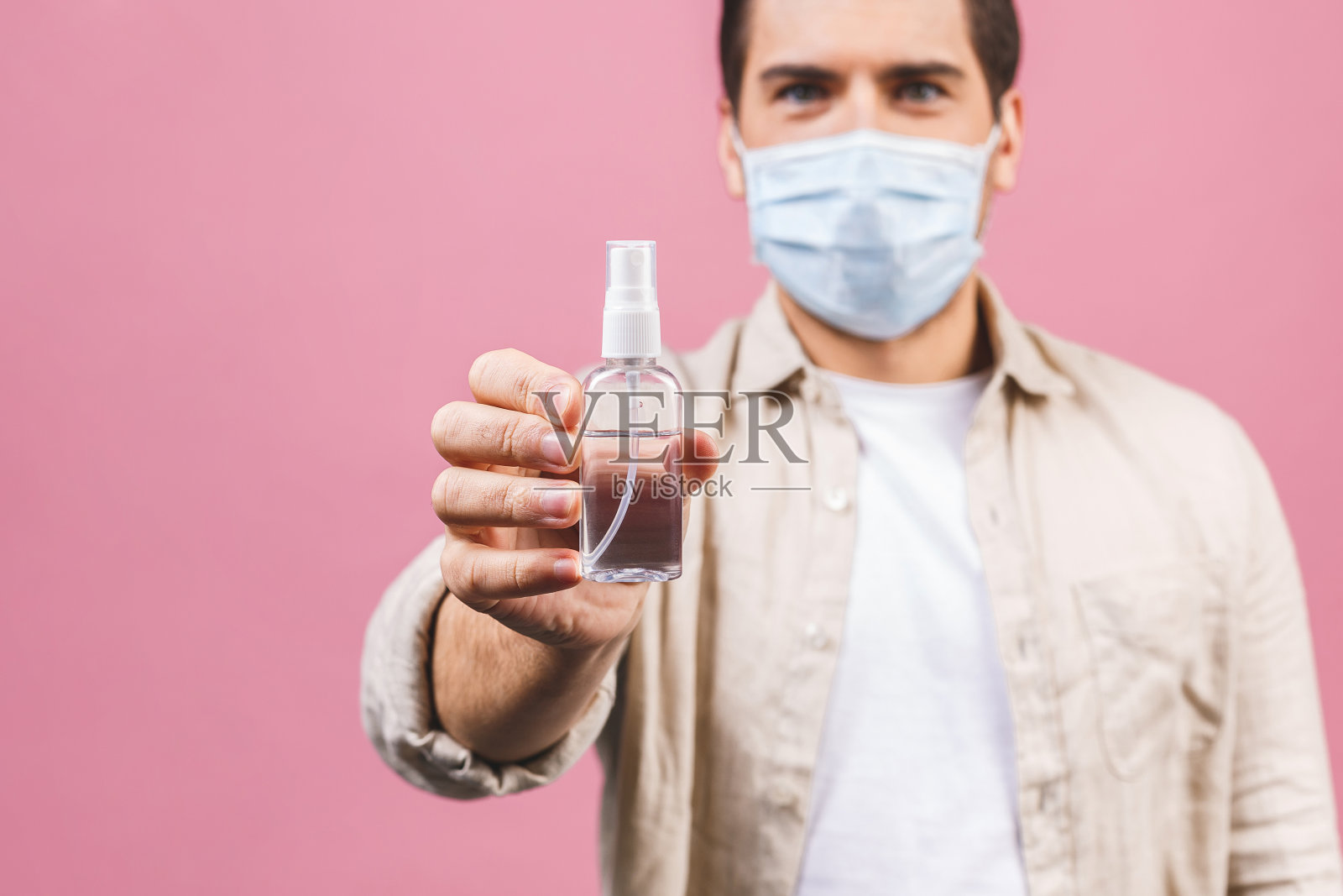 年轻男子戴无菌面罩，粉红色背景隔离。流行性冠状病毒2019-ncov sars covid-19流感病毒概念。装有酒精液体抗菌消毒剂的瓶子。照片摄影图片