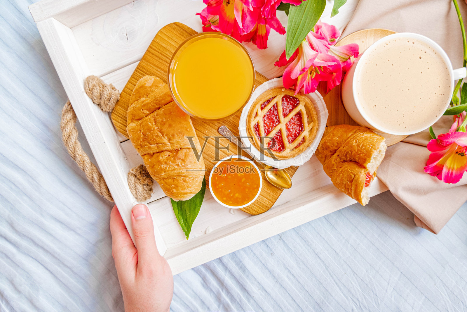 早餐在床上。一个男人端着早餐托盘，上面放着羊角面包、鲜花、咖啡、卡布奇诺、果酱、果汁和羊角面包。蓝色的彩色亚麻布。早上意外。情人节。度蜜月照片摄影图片