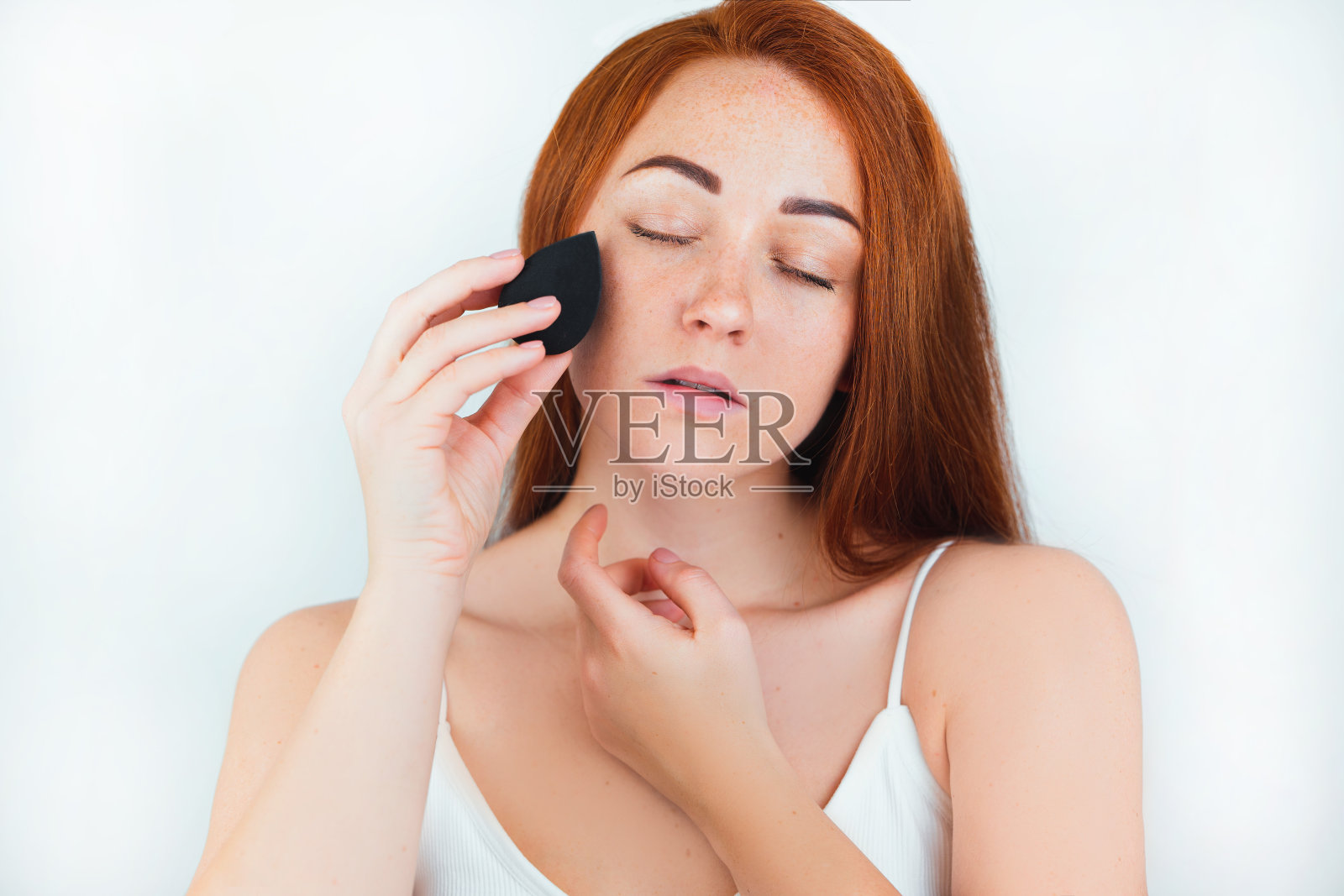 肖像红发年轻女子用化妆海绵在孤立的白色背景上涂抹皮肤粉底，护理和美丽的概念照片摄影图片