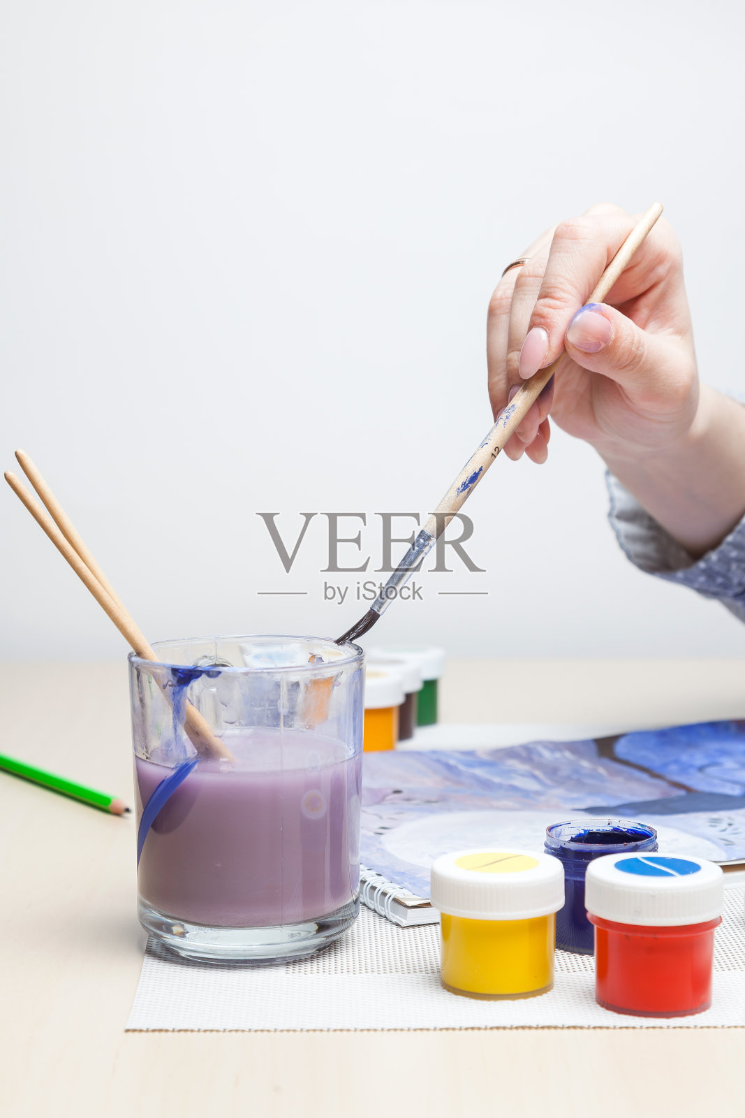 这是在艺术治疗工作室拍摄的一幅垂直镜头，一位女性的手拿着一支画笔，将它浸在一杯水里，水彩画的特写镜头被溶解在颜料罐的背景中。照片摄影图片