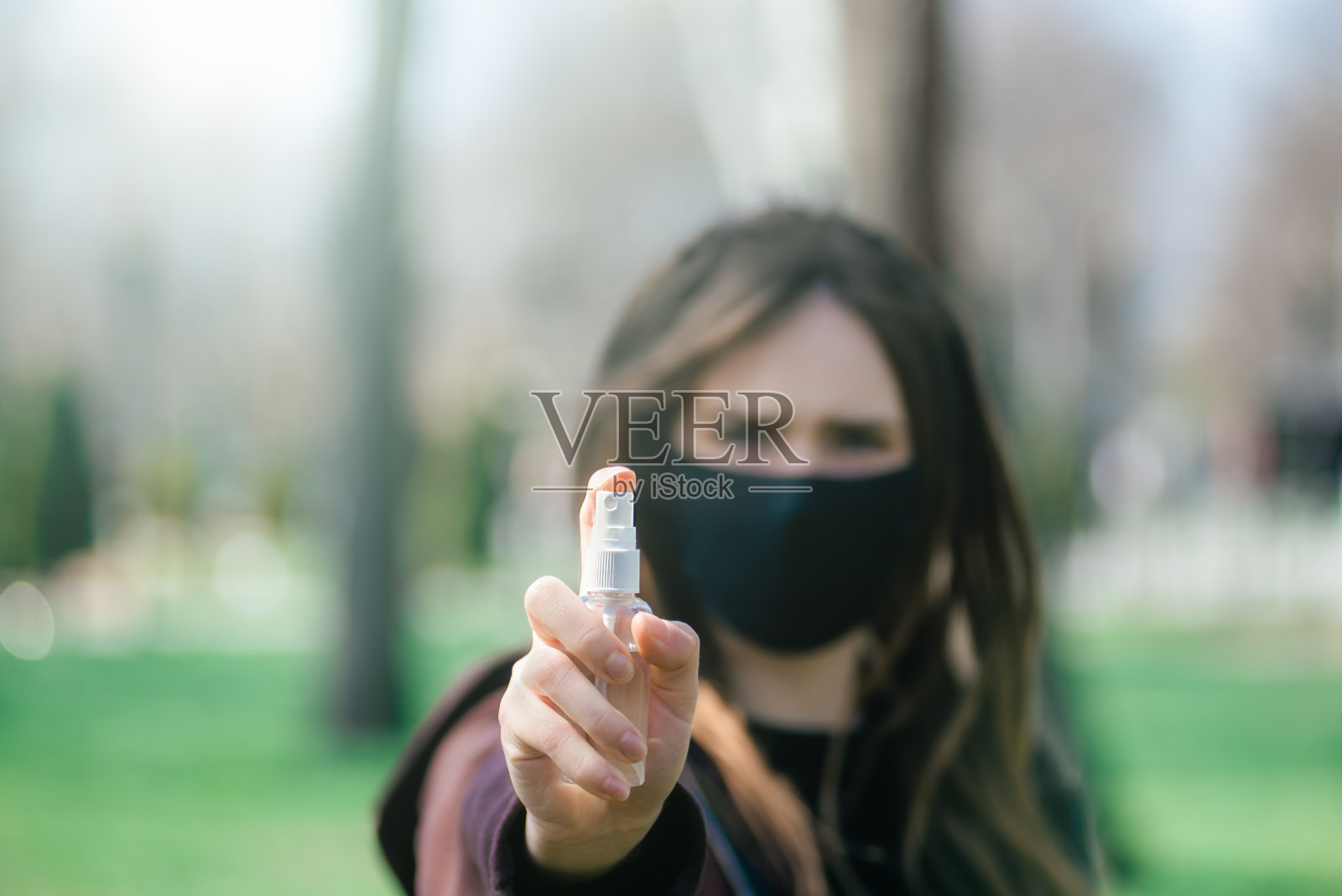 女孩将洗手液放在伸出的手臂上，重点是喷雾。一个带着医用防护面具的长发绺女孩的肖像。戴眼镜戴兜帽的女孩。照片摄影图片